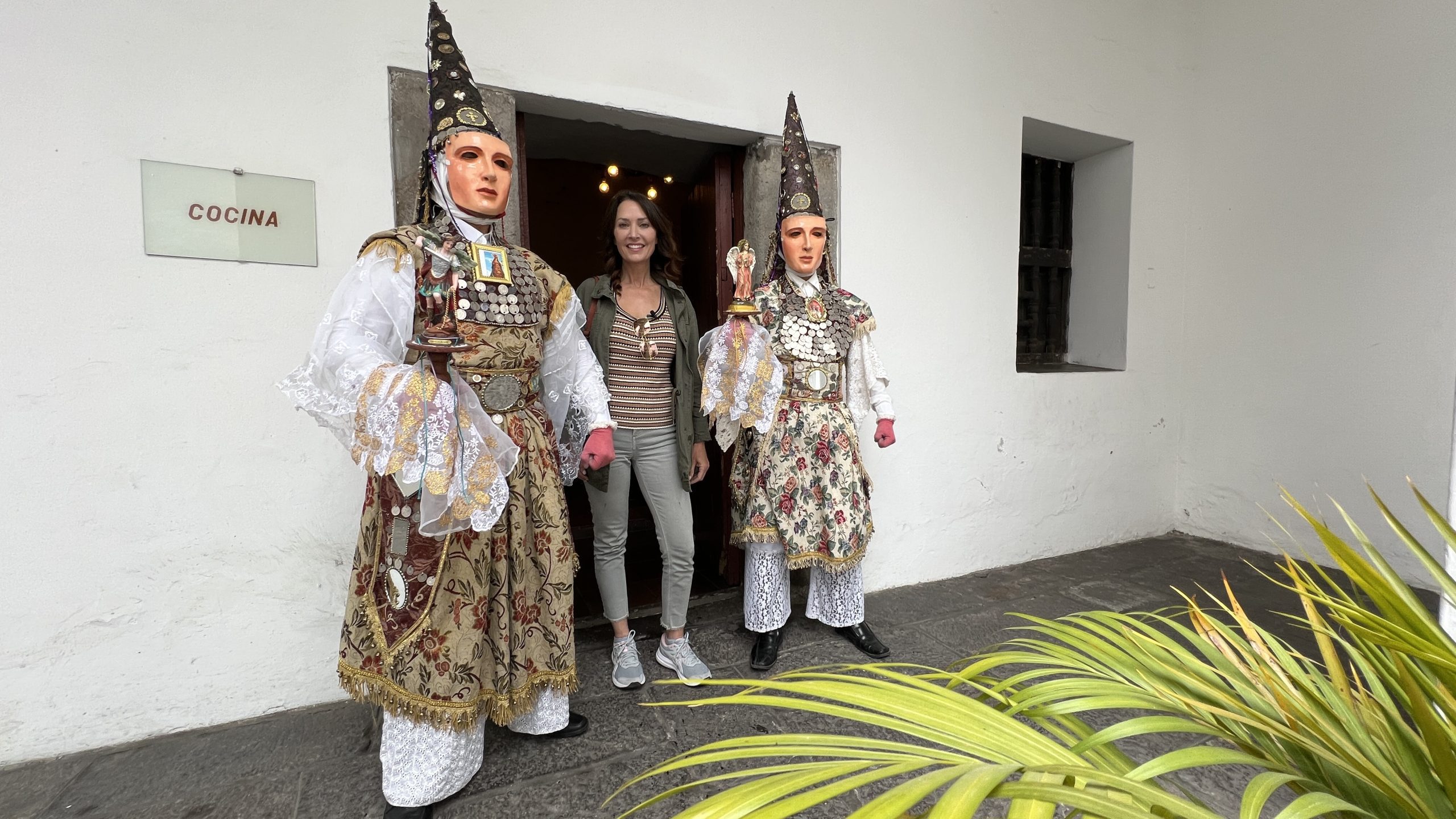 Two Danzantes de Quito dancers in mirrored cone hats and floral frocks beside a convent kitchen door