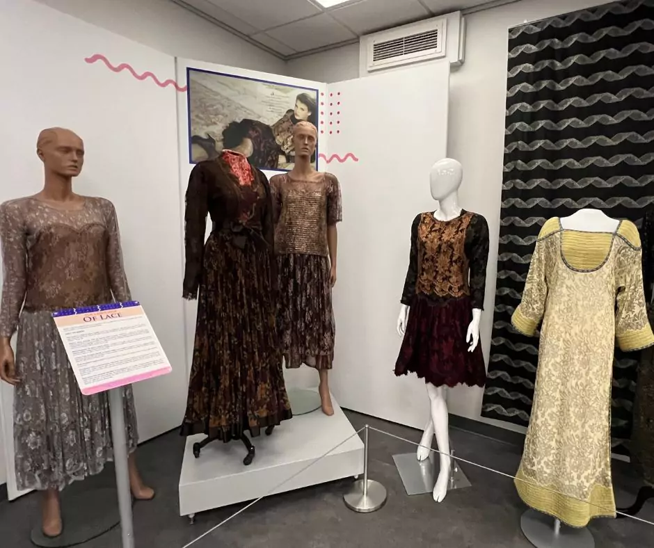 A Geoffrey Beene exhibit featuring mannequins dressed in intricate lace and brocade evening gowns in earthy tones. Informational signage titled “The Language of Lace” details Beene’s use of luxurious textiles.