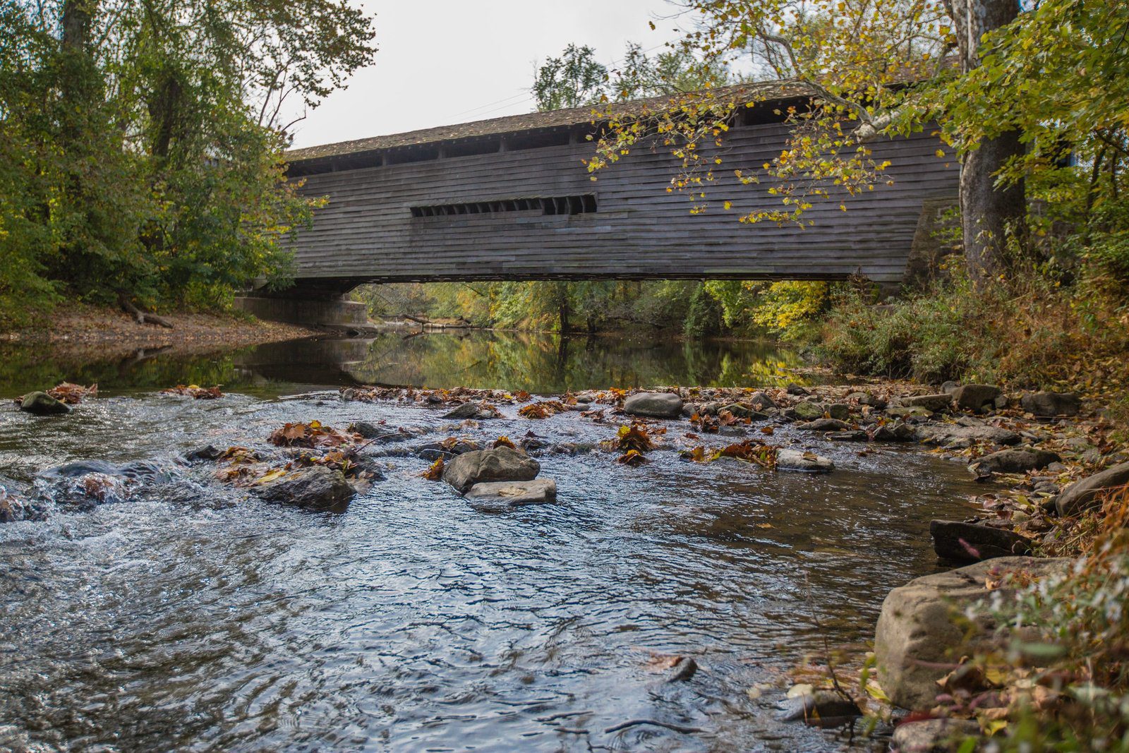 Dark wooden covered bridge crossing a calm rocky creek in a forested area during early fall, viewed from stream level.