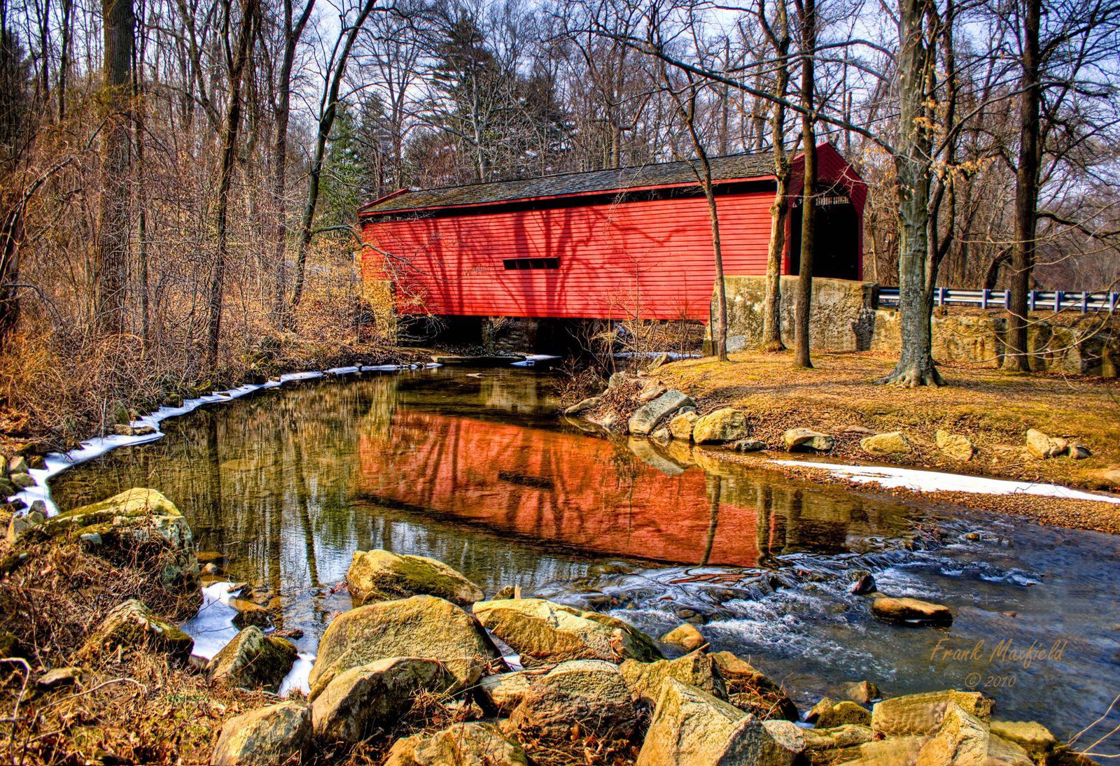 Red covered bridge crossing a reflective creek surrounded by bare trees and light snow, with golden afternoon light and early winter colors.