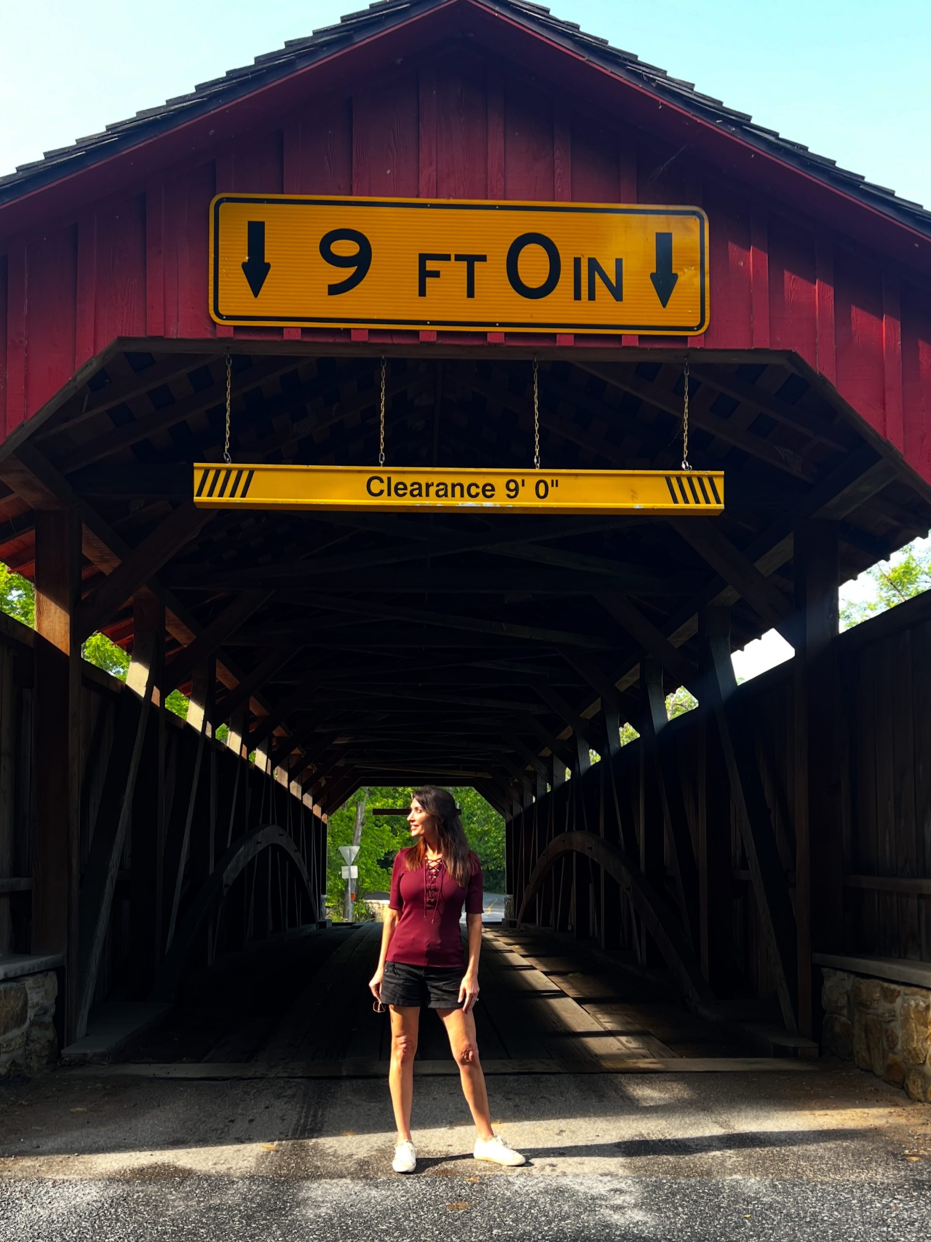 A woman poses under the roof of a red covered bridge, centered beneath a clearance sign and surrounded by natural light and wooden beams.