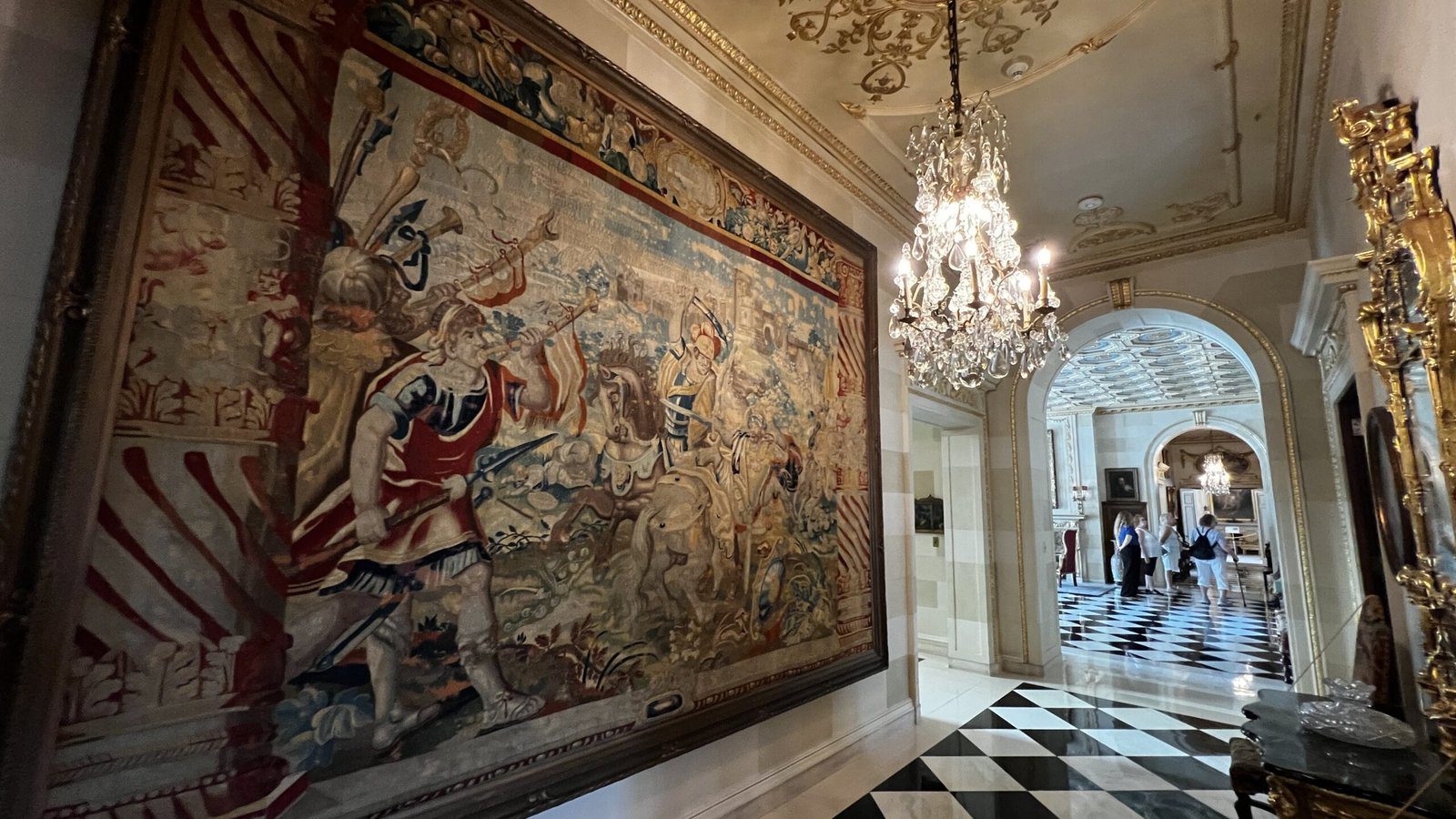 Elaborate hallway in Nemours Estate with a historic tapestry, chandeliers, and black-and-white marble floor
