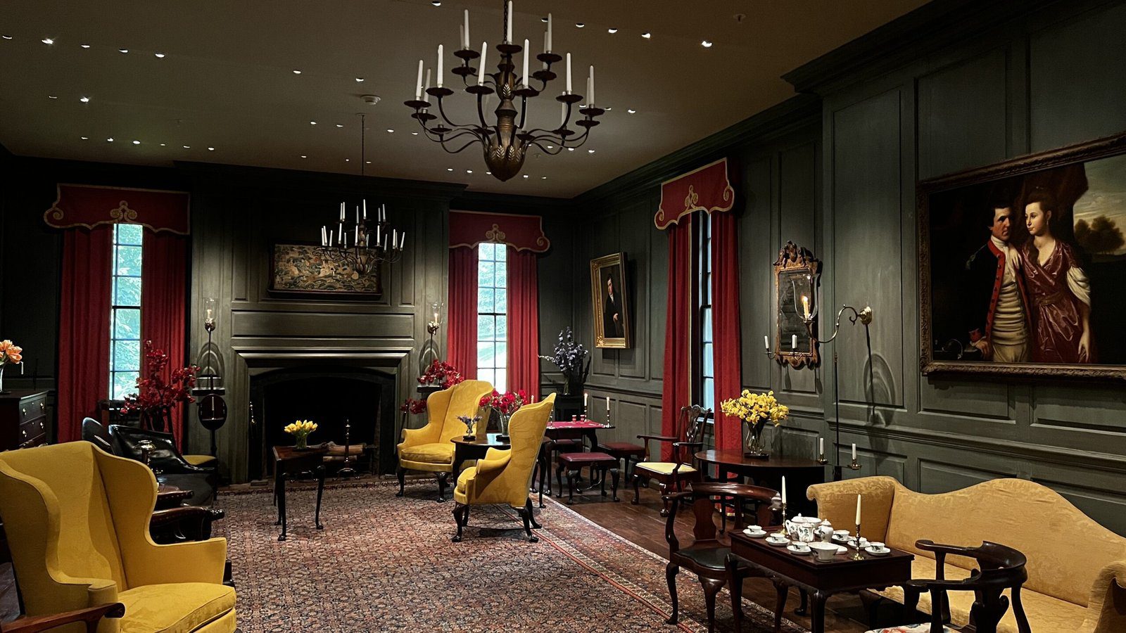 Warmly lit parlor at Winterthur with yellow armchairs, chandeliers, and colonial portraits lining the walls