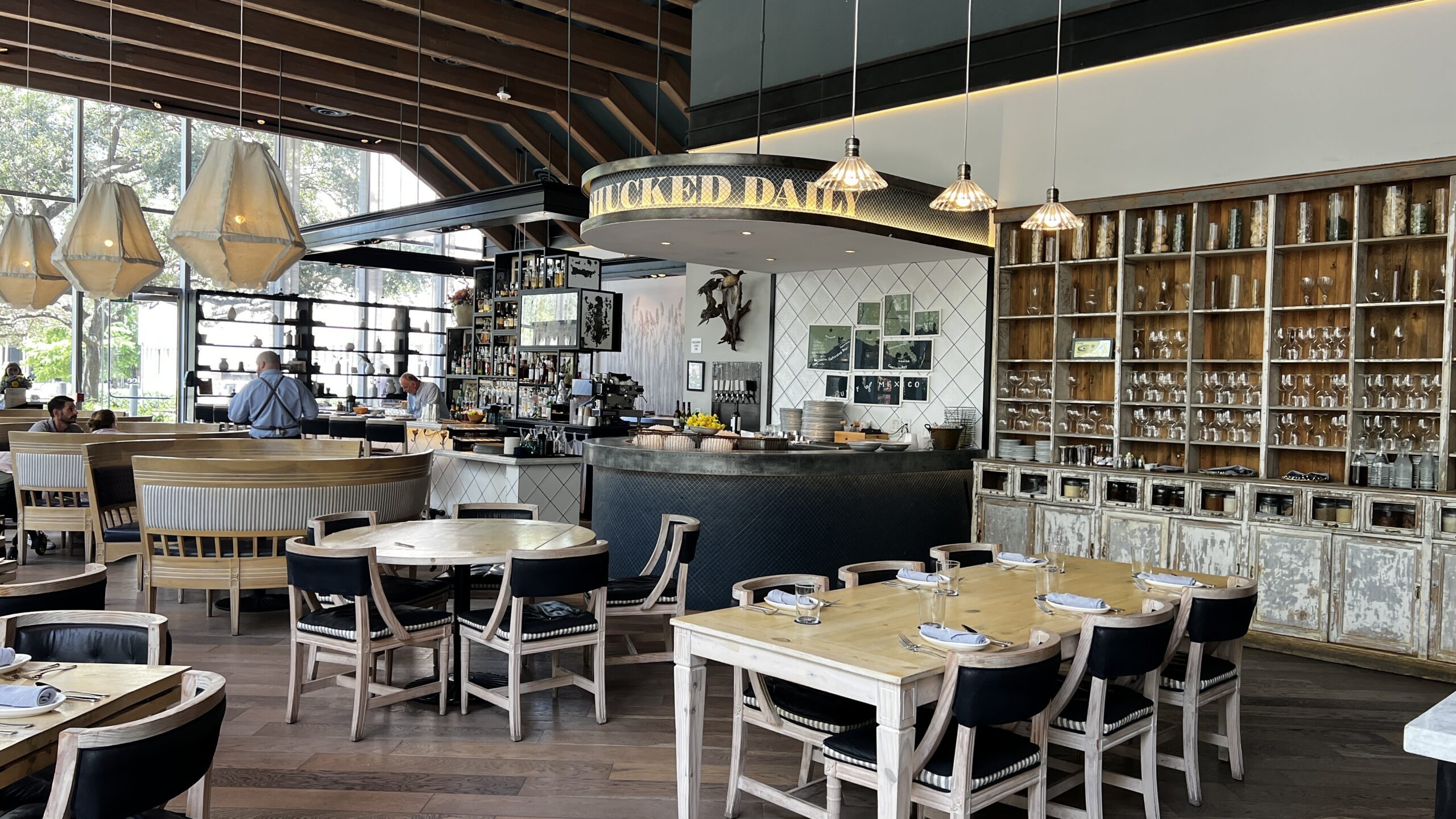 Bright dining room at Eunice in Houston featuring wood tables, modern lighting, and a raw bar with a “Shucked Daily” oyster counter.