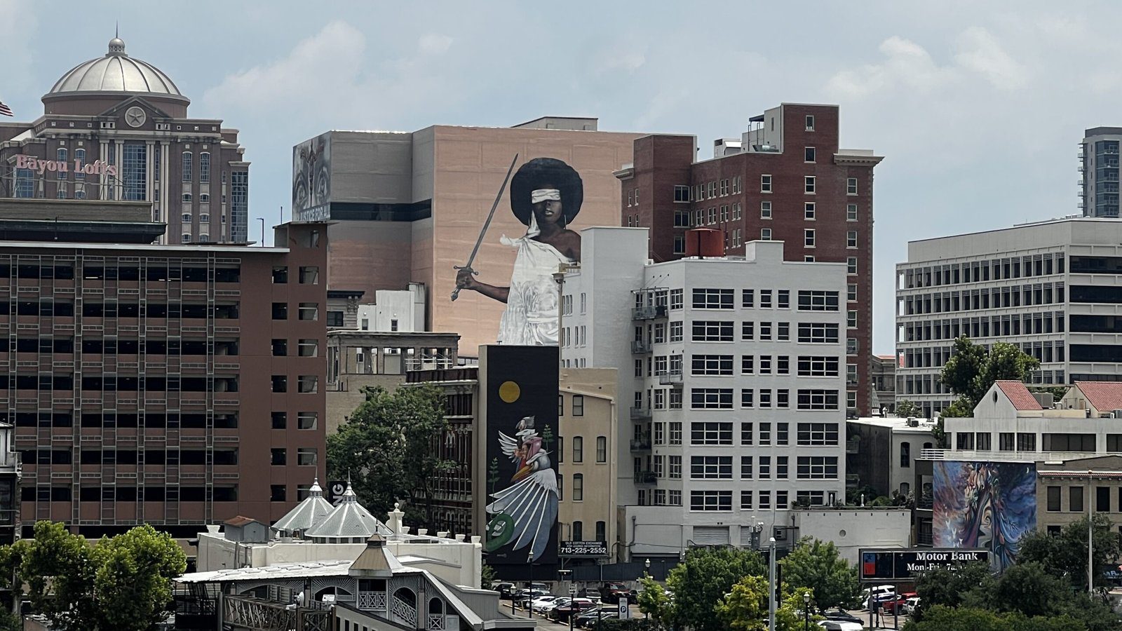 Tall mural of Lady Justice holding scales and a sword on the side of a downtown Houston building, surrounded by historic and modern architecture.