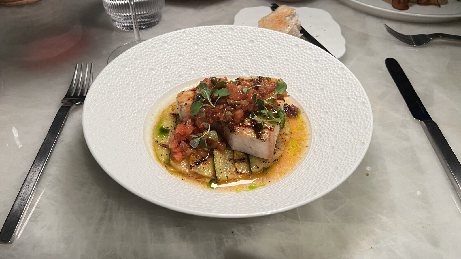 White plate of seared fish fillet with zucchini, tomato compote, and herb garnish at Le Jardinier in Houston.