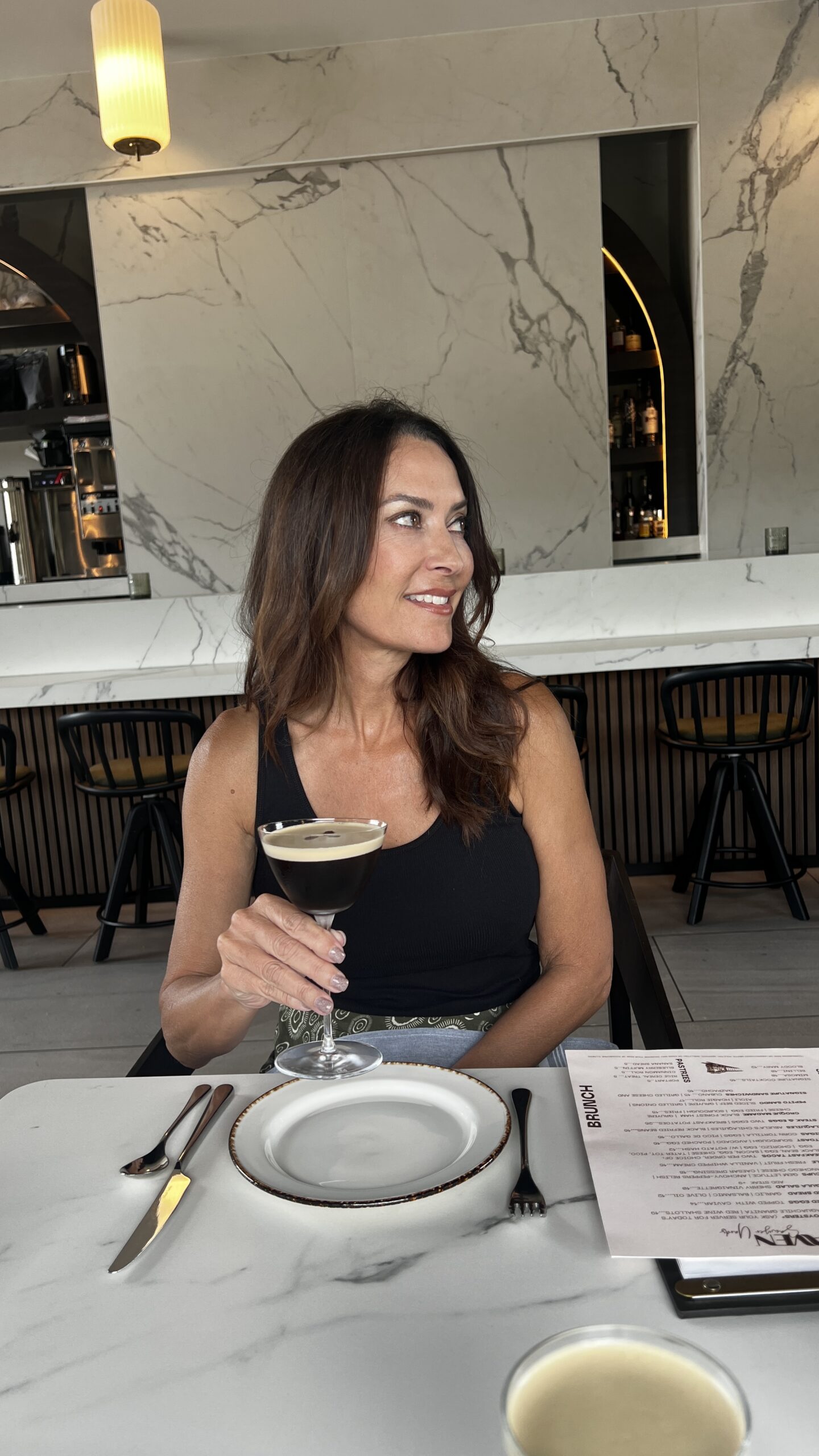 Woman enjoying an espresso martini at Maven in Houston, seated at a marble table with modern bar design in the background.