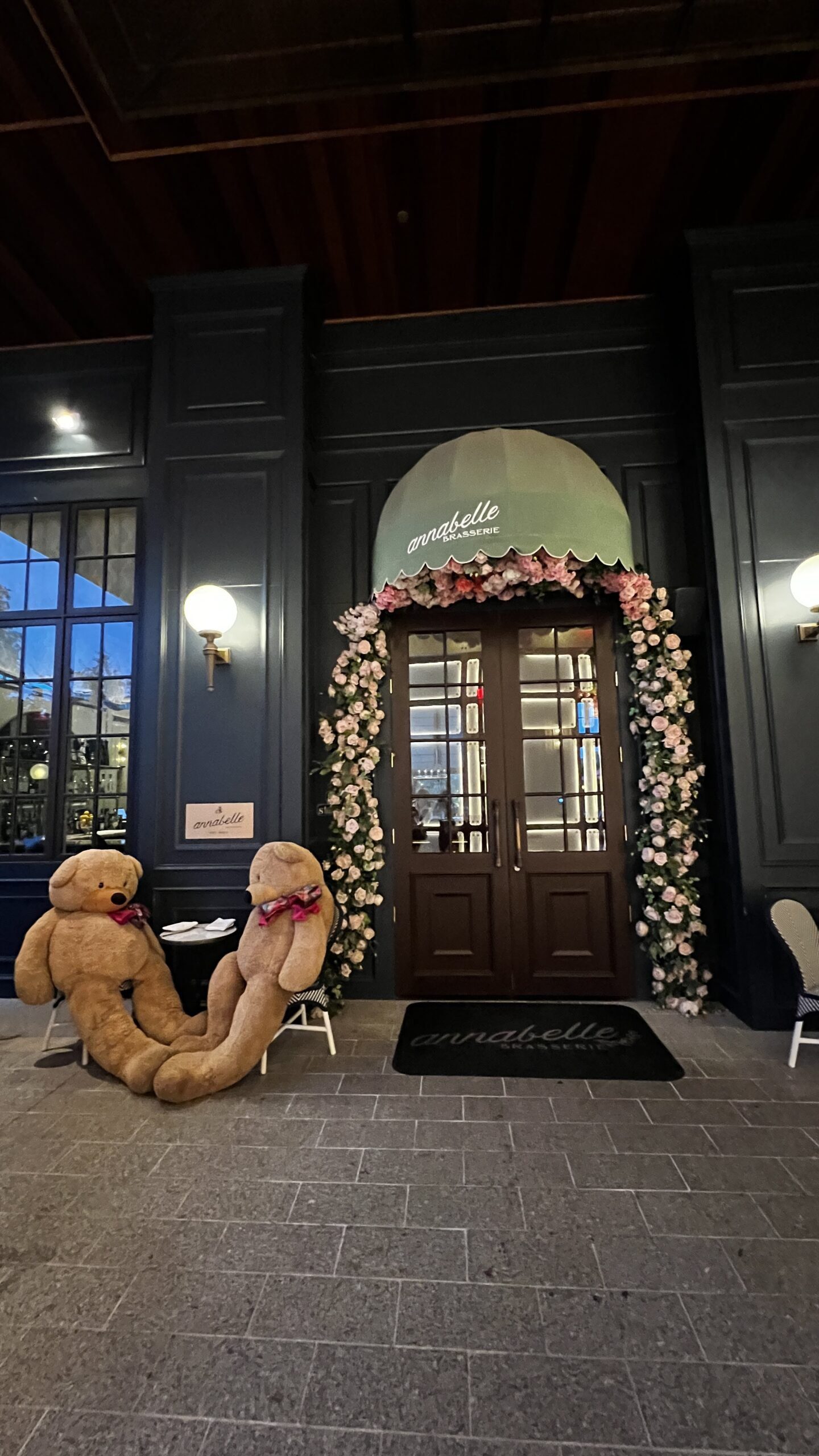 Two oversized teddy bears seated beside the floral-framed entrance of Annabelle Brasserie in Houston.