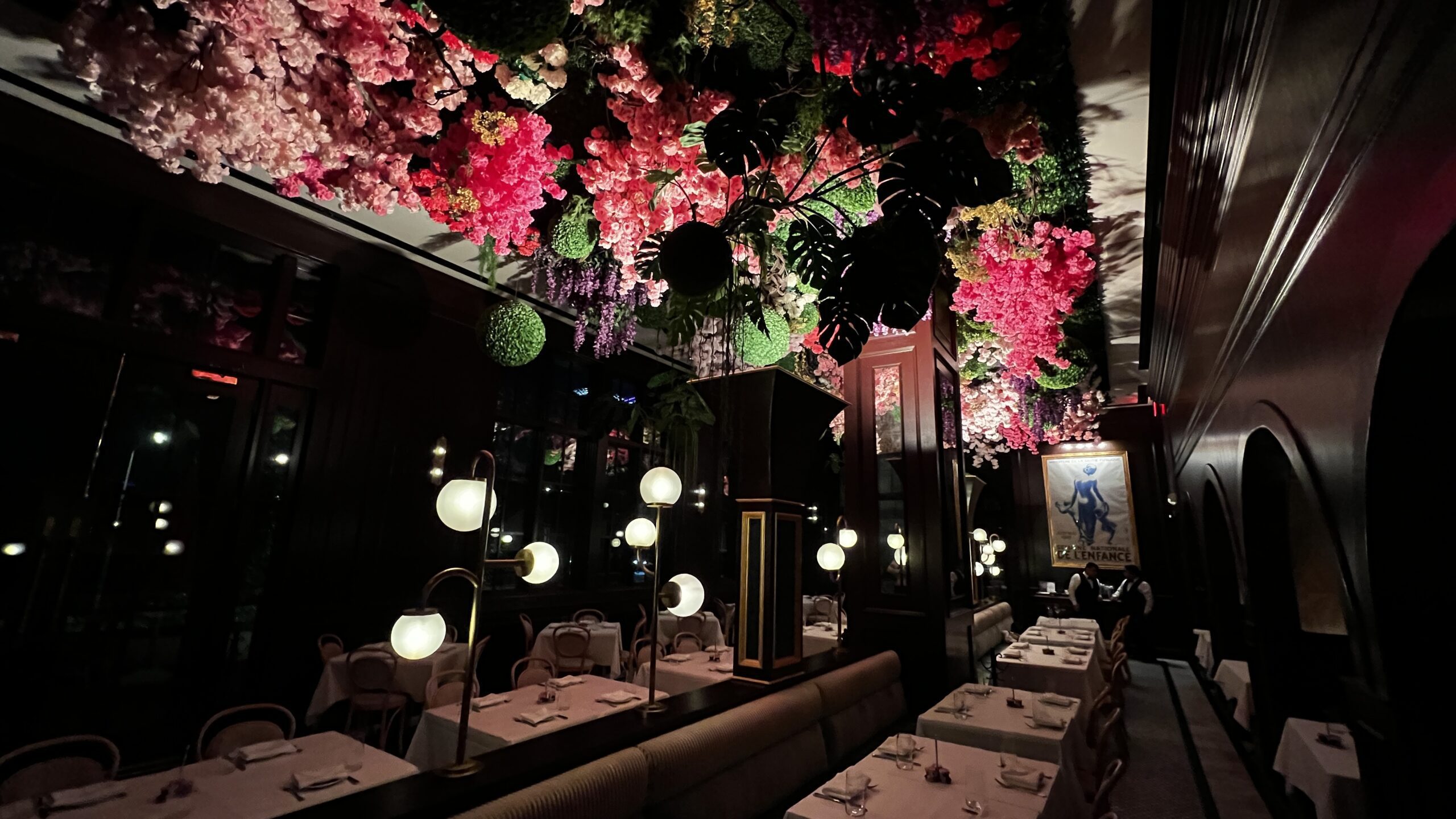 Elegant restaurant interior with white tablecloth dining tables, globe pendant lamps, and a dramatic ceiling installation of lush greenery and cascading pink, red, and purple flowers.