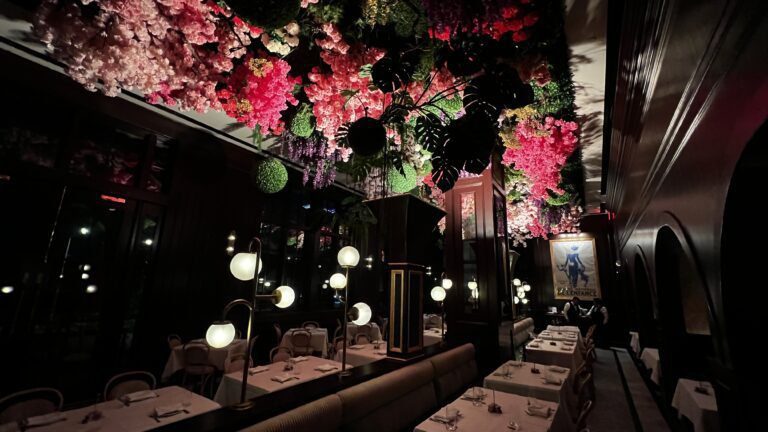 Elegant restaurant interior with white tablecloth dining tables, globe pendant lamps, and a dramatic ceiling installation of lush greenery and cascading pink, red, and purple flowers - experiencing culinary culture in Bayou City.
