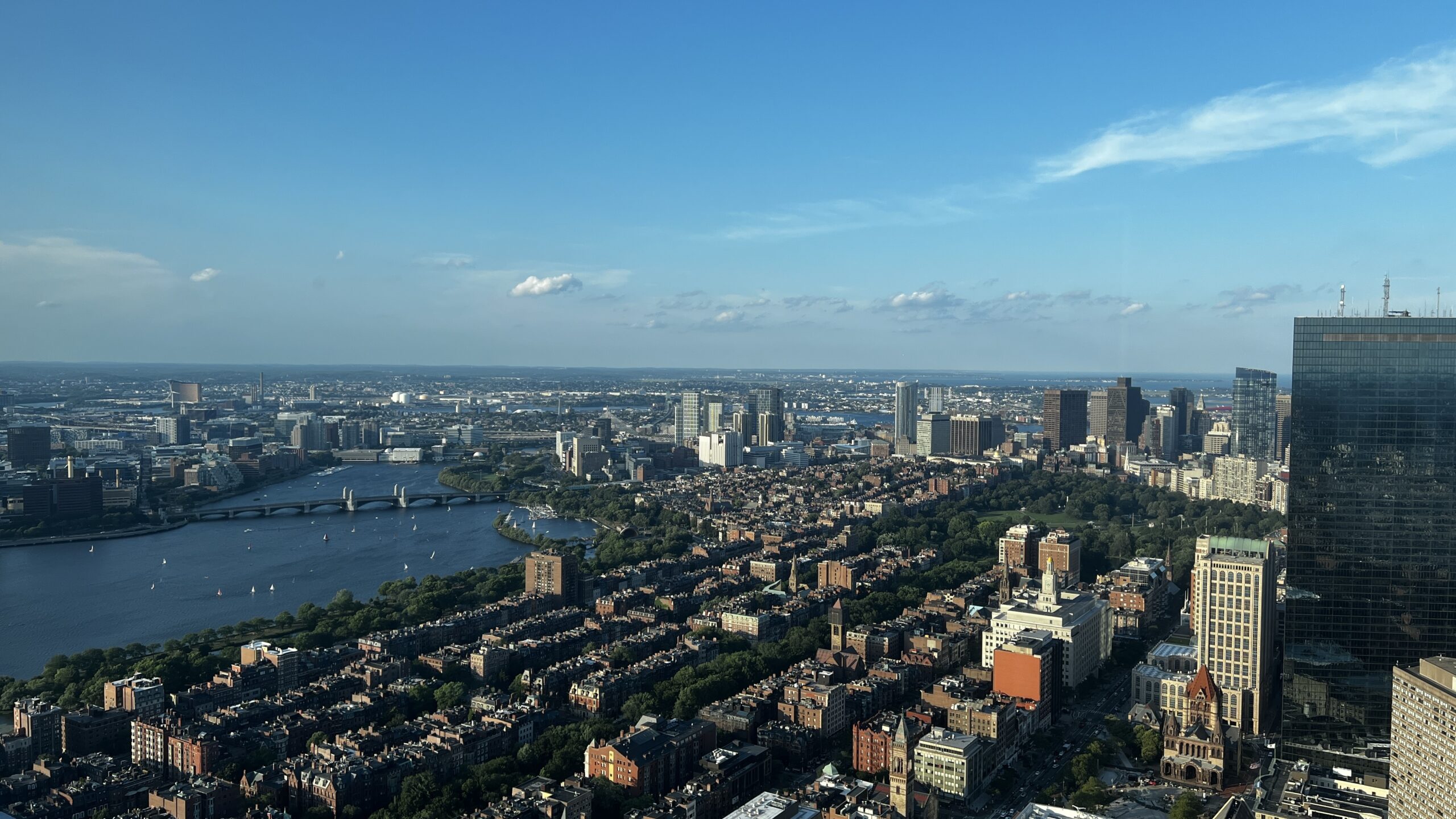 Aerial view of Boston showing the Charles River, Back Bay neighborhood with brownstone rowhouses, and downtown skyline under blue sky