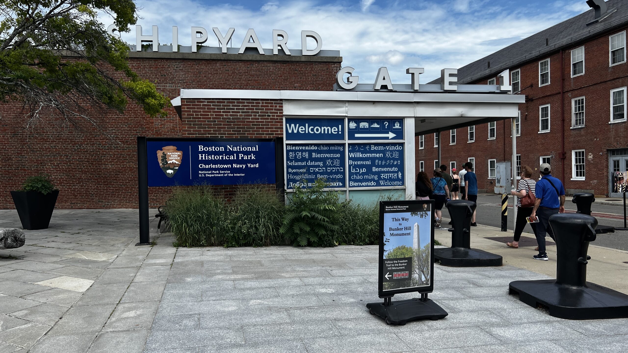 Entrance to Charlestown Navy Yard showing brick buildings with 'Shipyard' and 'Gate 1' signage, National Park Service sign, visitors walking through plaza