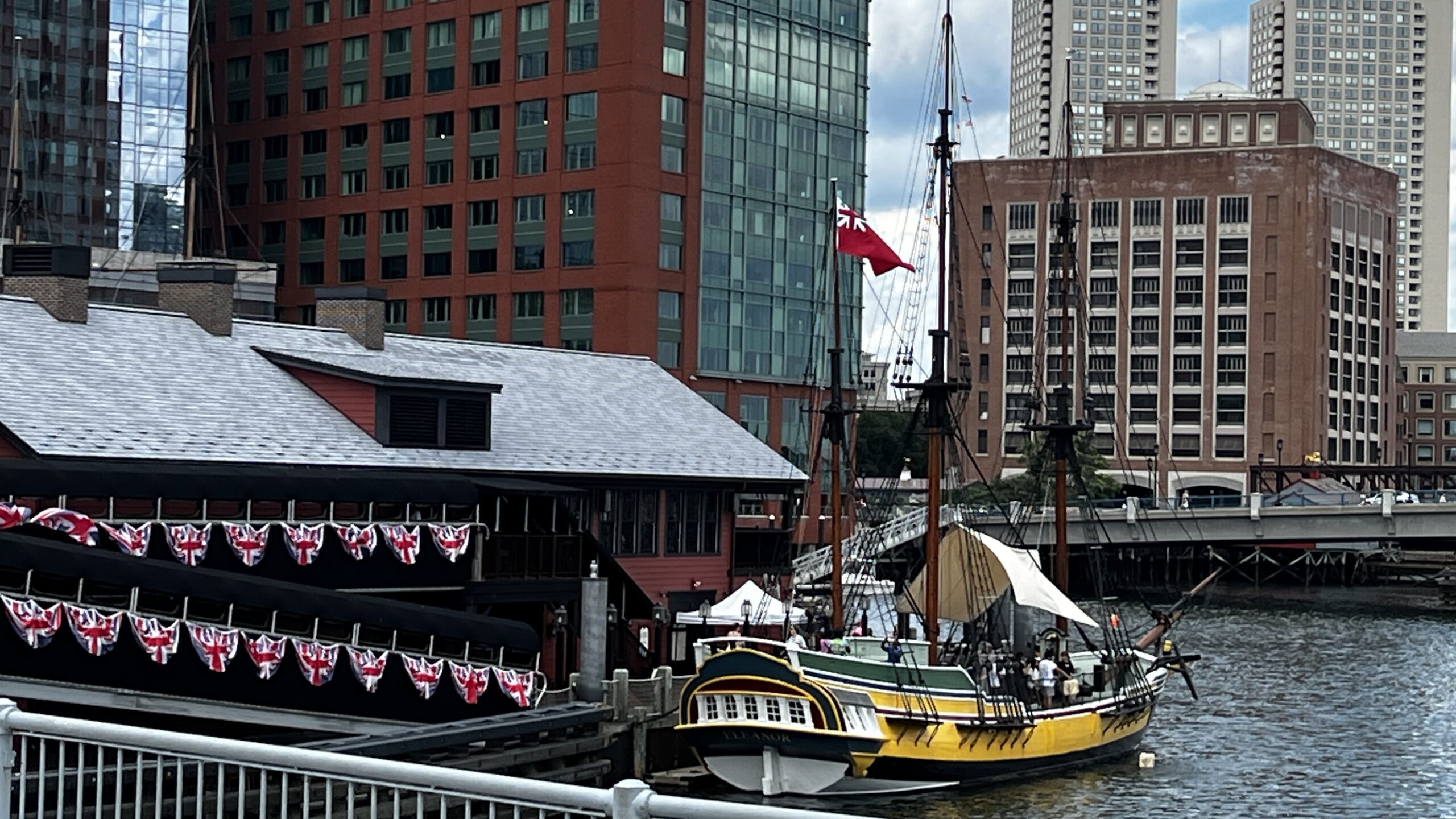 Replica 18th-century sailing ship with yellow and black hull docked at Boston Tea Party Museum, modern city buildings in background, patriotic bunting displayed