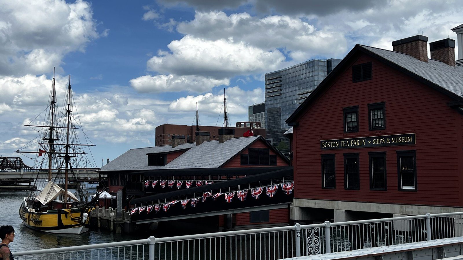 Historic red wooden building labeled 'Boston Tea Party Ships & Museum' with replica colonial ship docked alongside, American flag bunting decoration, waterfront setting