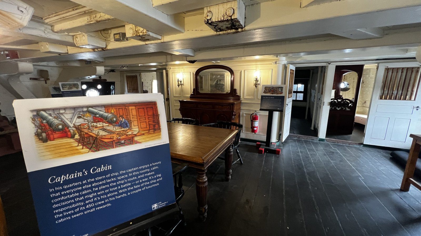 Interior of ship's captain's cabin showing dark wooden floors, white walls, period furniture including large wooden table, fireplace mantel, and interpretive sign