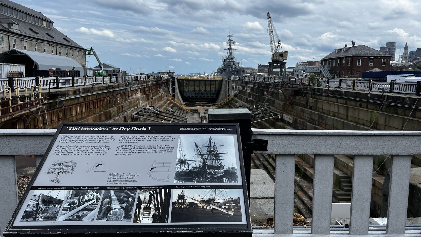 Empty historic dry dock with steep walls and cranes visible, interpretive sign in foreground explaining 'Old Ironsides in Dry Dock 1' with historical photos