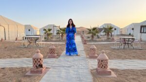 Woman in a blue patterned dress standing on a walkway lined with lanterns at a Sahara desert camp surrounded by white tents and palm trees.