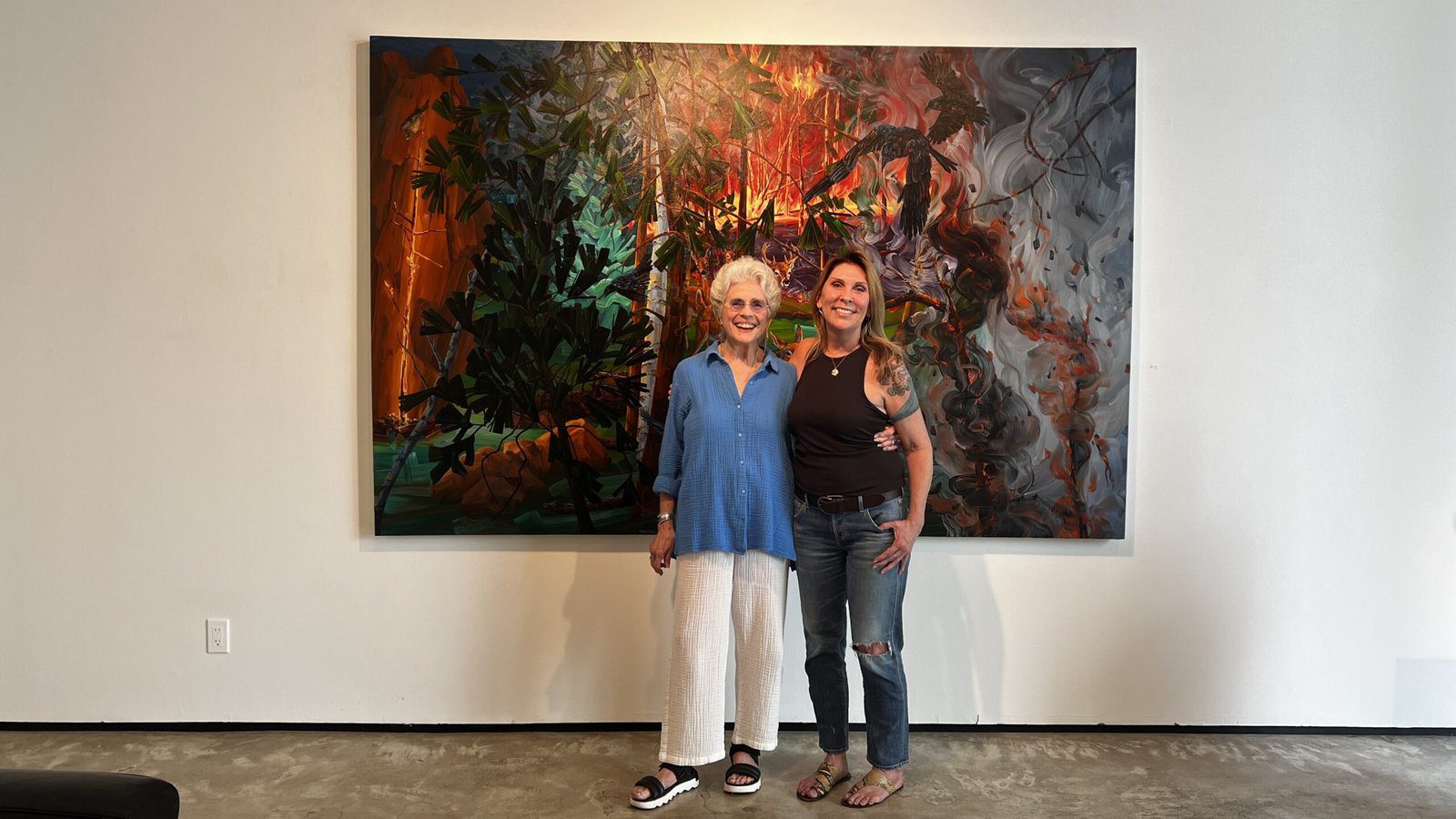 Two women standing together in front of a large abstract forest-inspired painting inside a Houston gallery.
