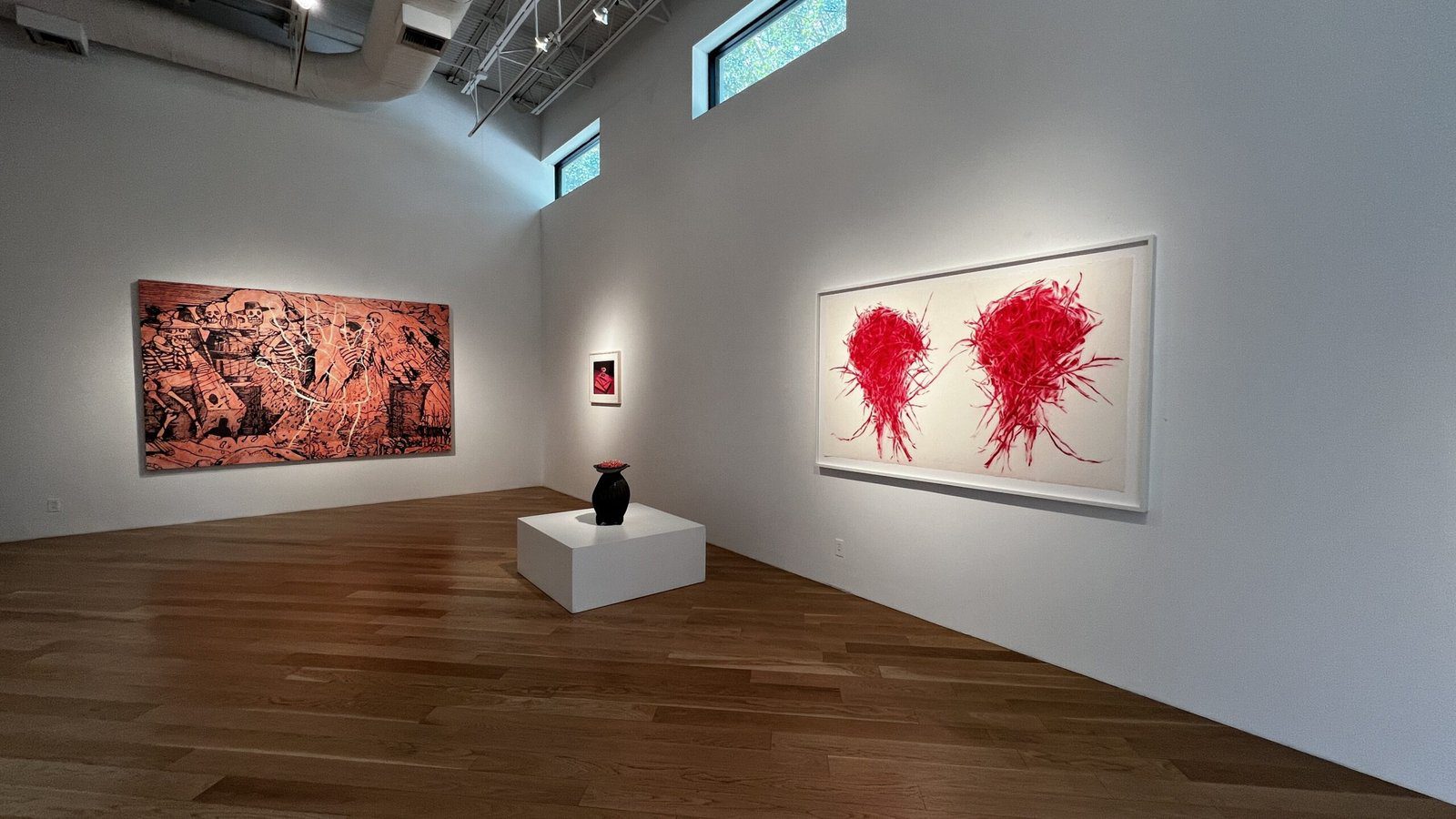 White-walled gallery with wooden floors featuring a large red abstract painting, a black vase on a pedestal, and contemporary artworks.