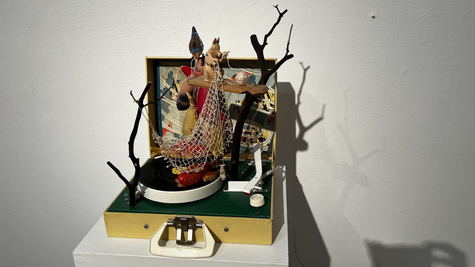 Mixed-media sculpture using a vintage record player, branches, netting, and figurines displayed on a white pedestal.