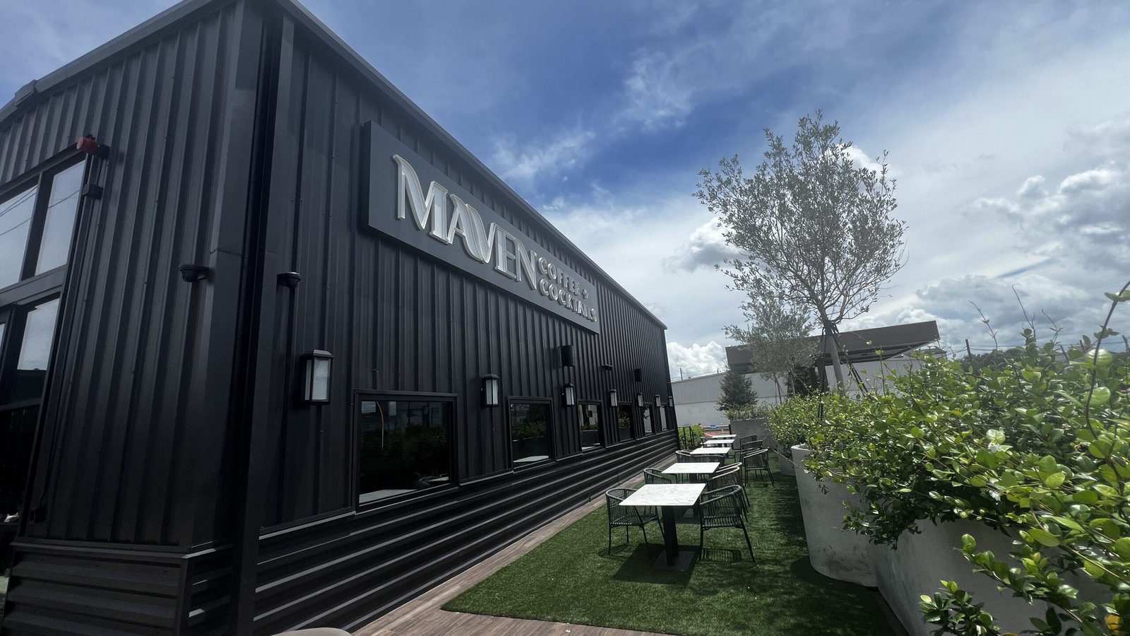 Black exterior of Maven Coffee and Cocktails with outdoor seating and greenery under a bright blue sky.