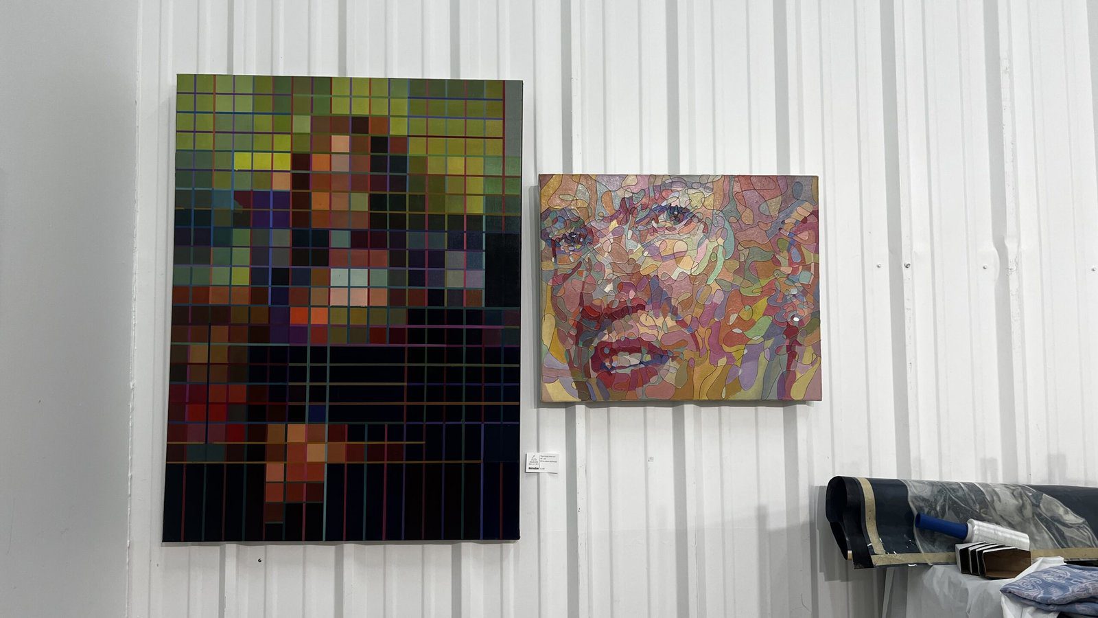 Two contemporary artworks on a gallery wall: a large pixelated grid-style portrait and a colorful mosaic portrait of a face.