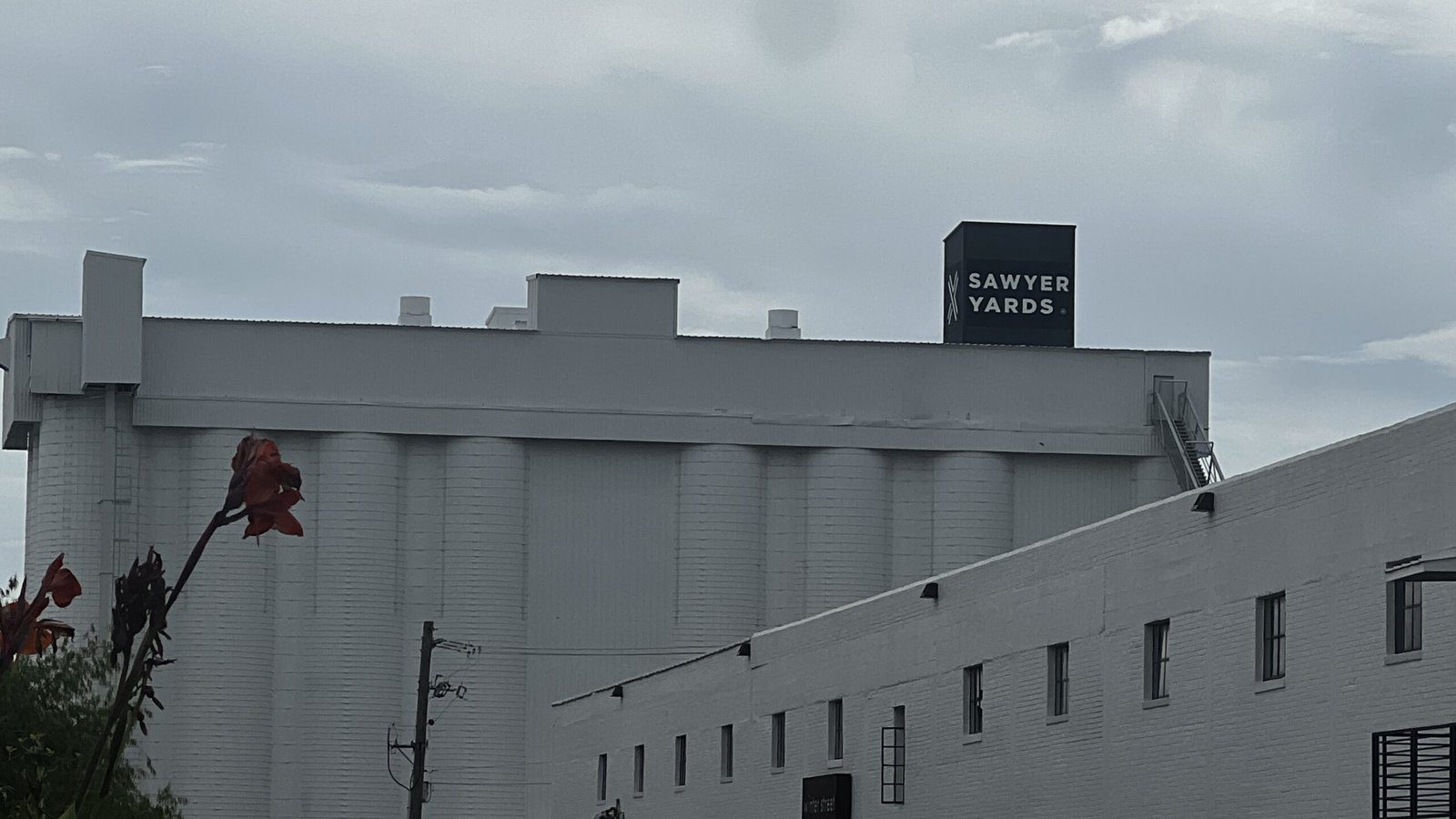 White industrial building with “Sawyer Yards” signage on a tall structure, seen under a cloudy sky.