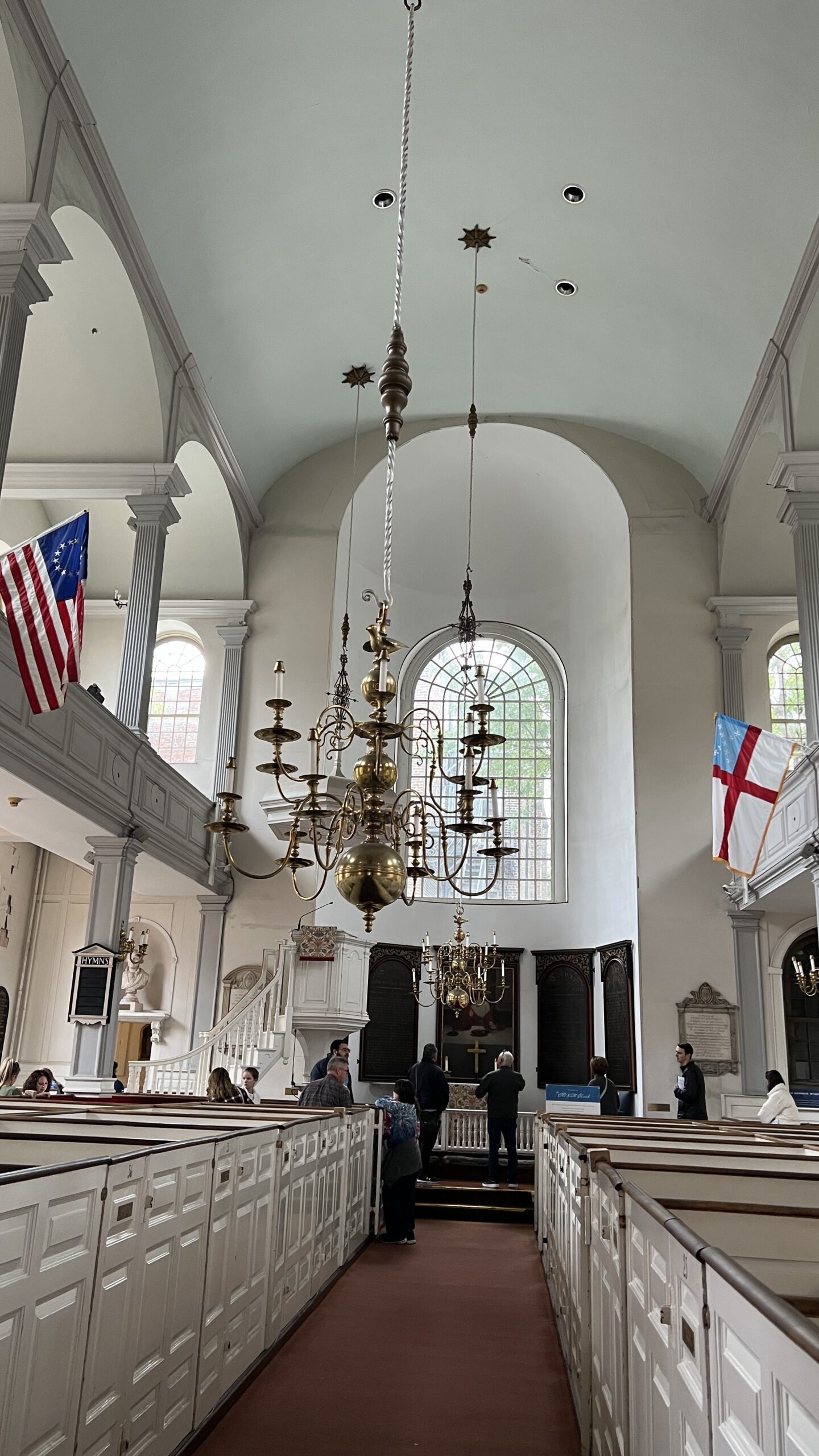 Interior of historic white church with vaulted ceiling, wooden box pews, ornate brass chandeliers, American and Episcopal flags, and arched windows