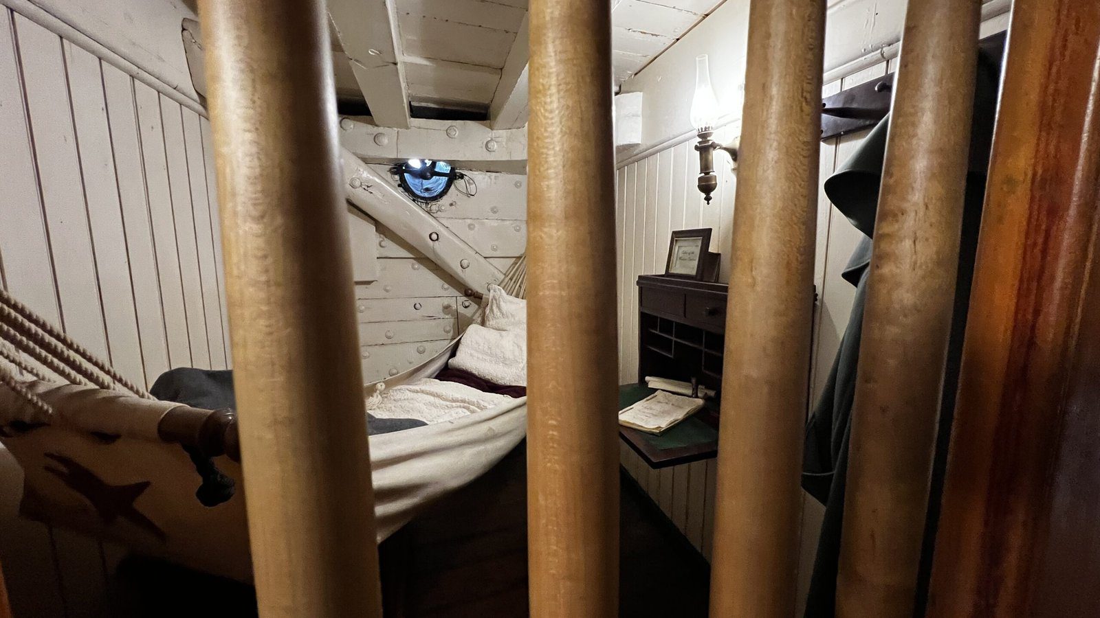 View through brass bars or railings into cramped ship quarters showing hammock, white wooden walls, and simple furnishings in dim lighting