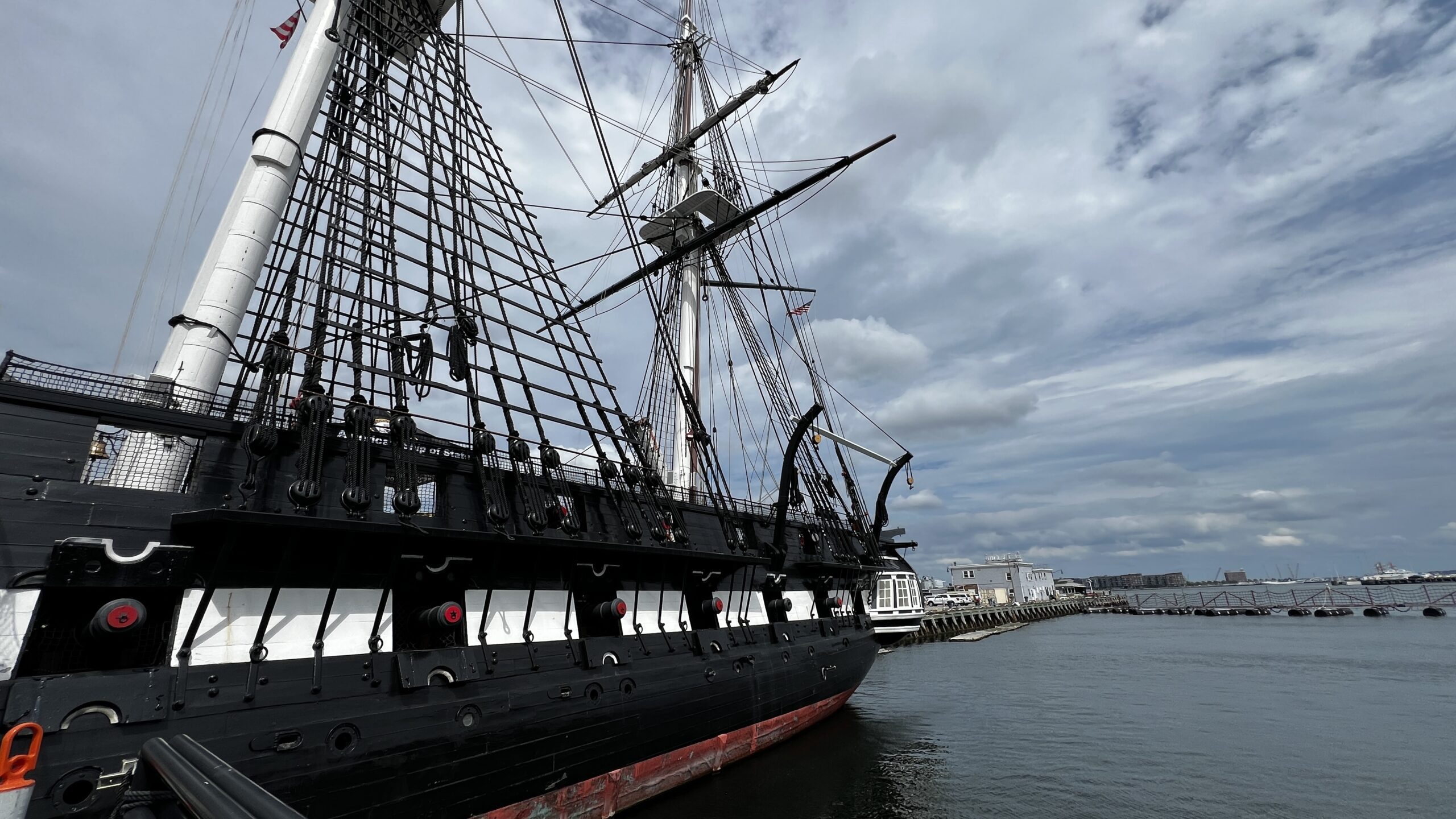Close-up side view of USS Constitution's black hull showing gun ports, complex rigging, rope ladders, and masts against cloudy sky and harbor backdrop