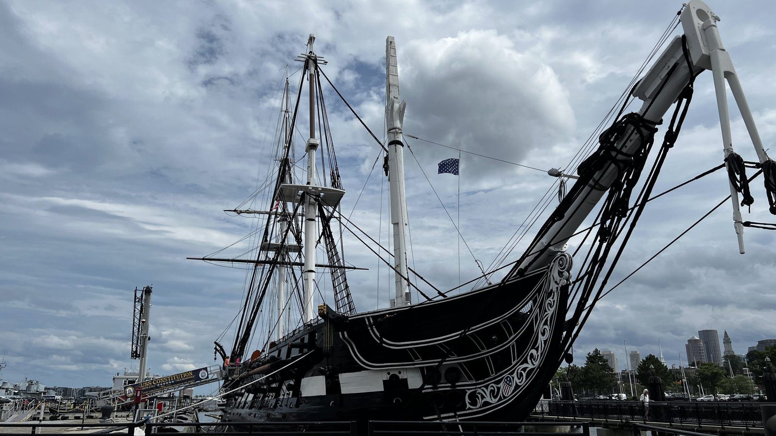 Side view of USS Constitution tall ship under dramatic cloudy sky, showing black hull with ornate carved stern and complex rigging of masts