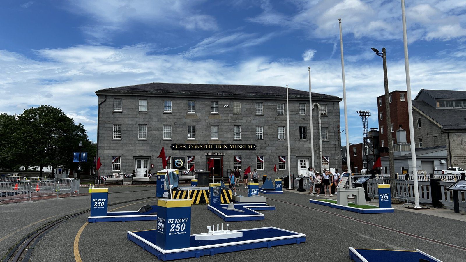 Gray three-story USS Constitution Museum building with rows of windows, flagpoles in front, and blue commemorative displays in foreground plaza