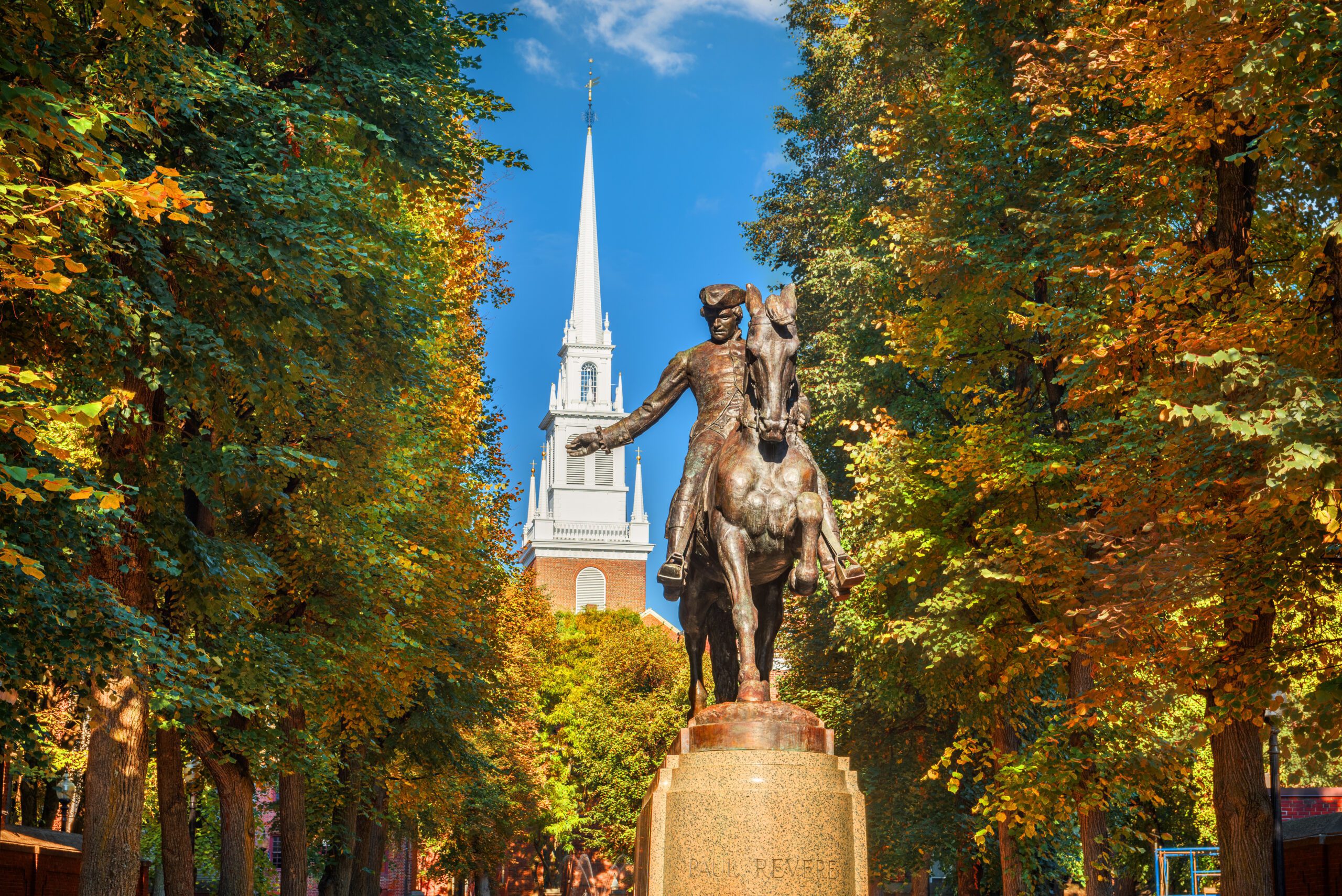 Bronze equestrian statue of Paul Revere on horseback framed by vibrant autumn foliage in orange, yellow, and green, with white steeple of Old North Church rising against blue sky in background