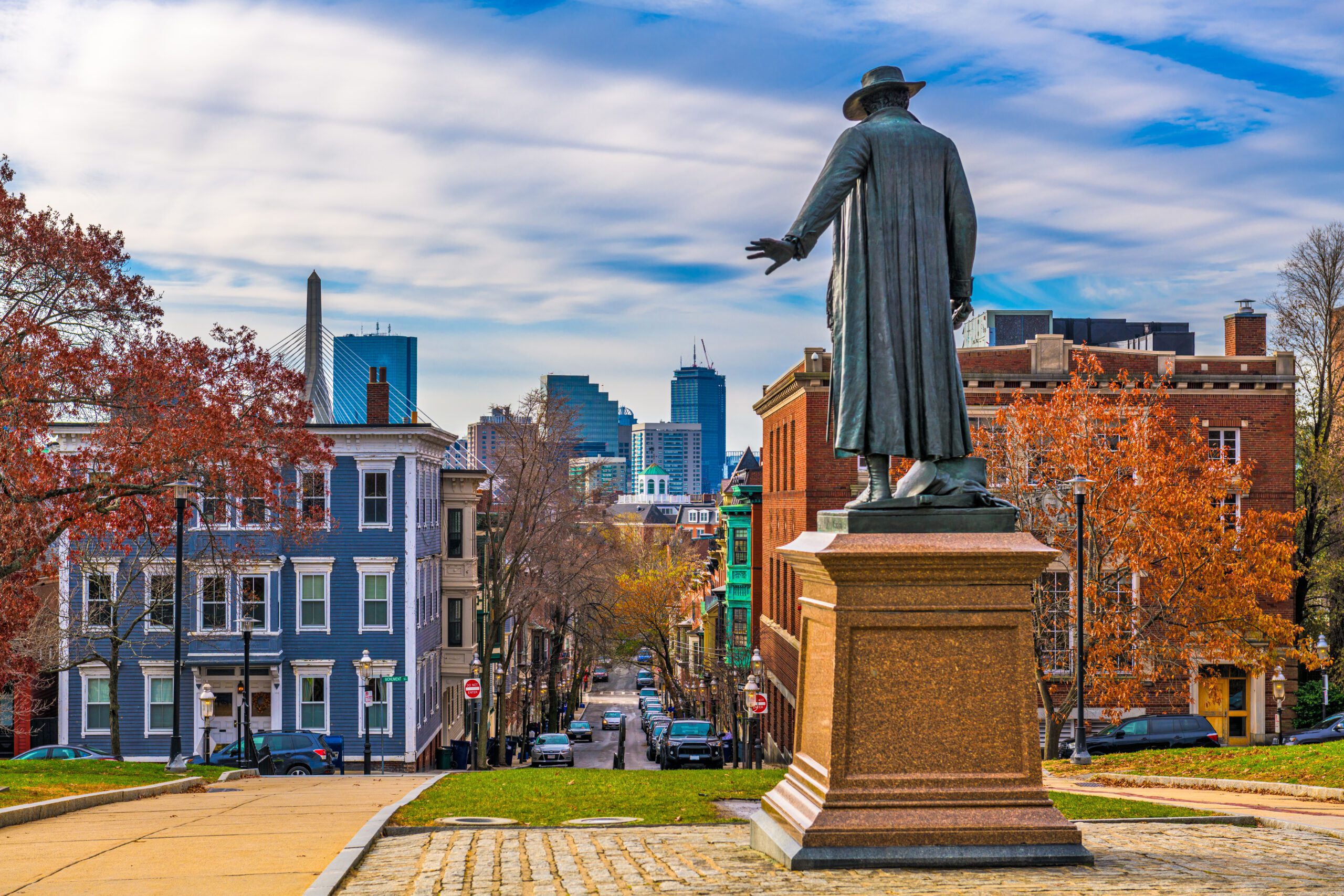 Bronze statue of colonial figure in long coat and hat standing on pedestal, viewed from behind, overlooking tree-lined street with historic rowhouses and modern Boston skyline in distance during autumn