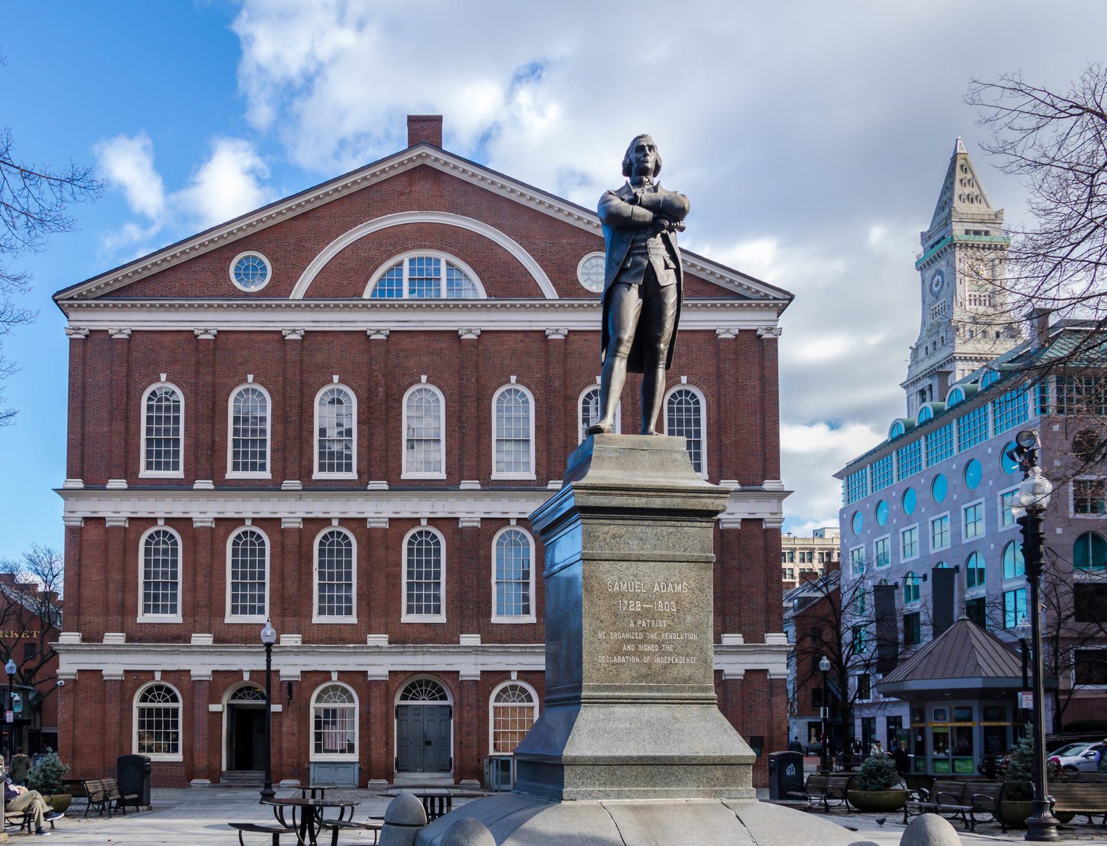 Bronze statue of Samuel Adams standing with arms crossed in front of Faneuil Hall's red brick colonial facade