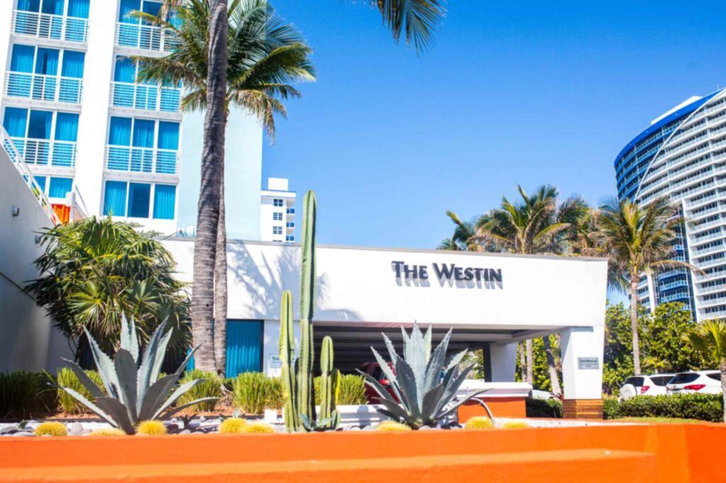 A bright daytime shot of The Westin hotel entrance portico under a clear blue sky. The white concrete structure features black text signage reading The Westin, flanked by tall palm trees and a landscaped garden bed with agave plants and a cactus behind a bright orange retaining wall.