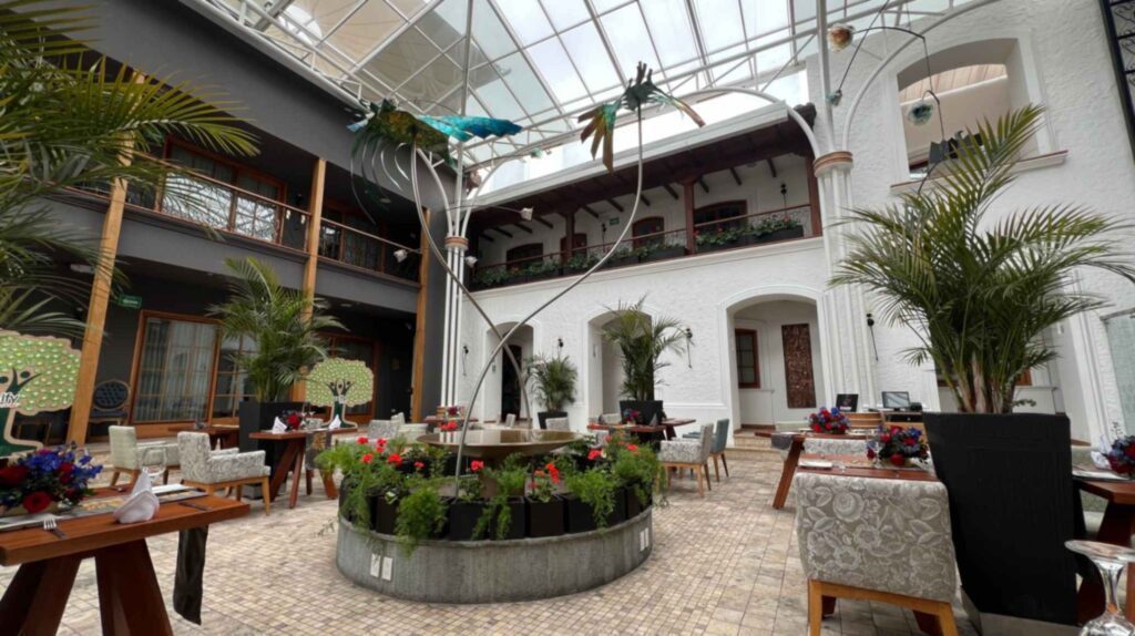 Eye level perspective of an indoor courtyard restaurant in Ecuador showing white stone arches wooden pillars potted palms and dining tables set with floral centerpieces