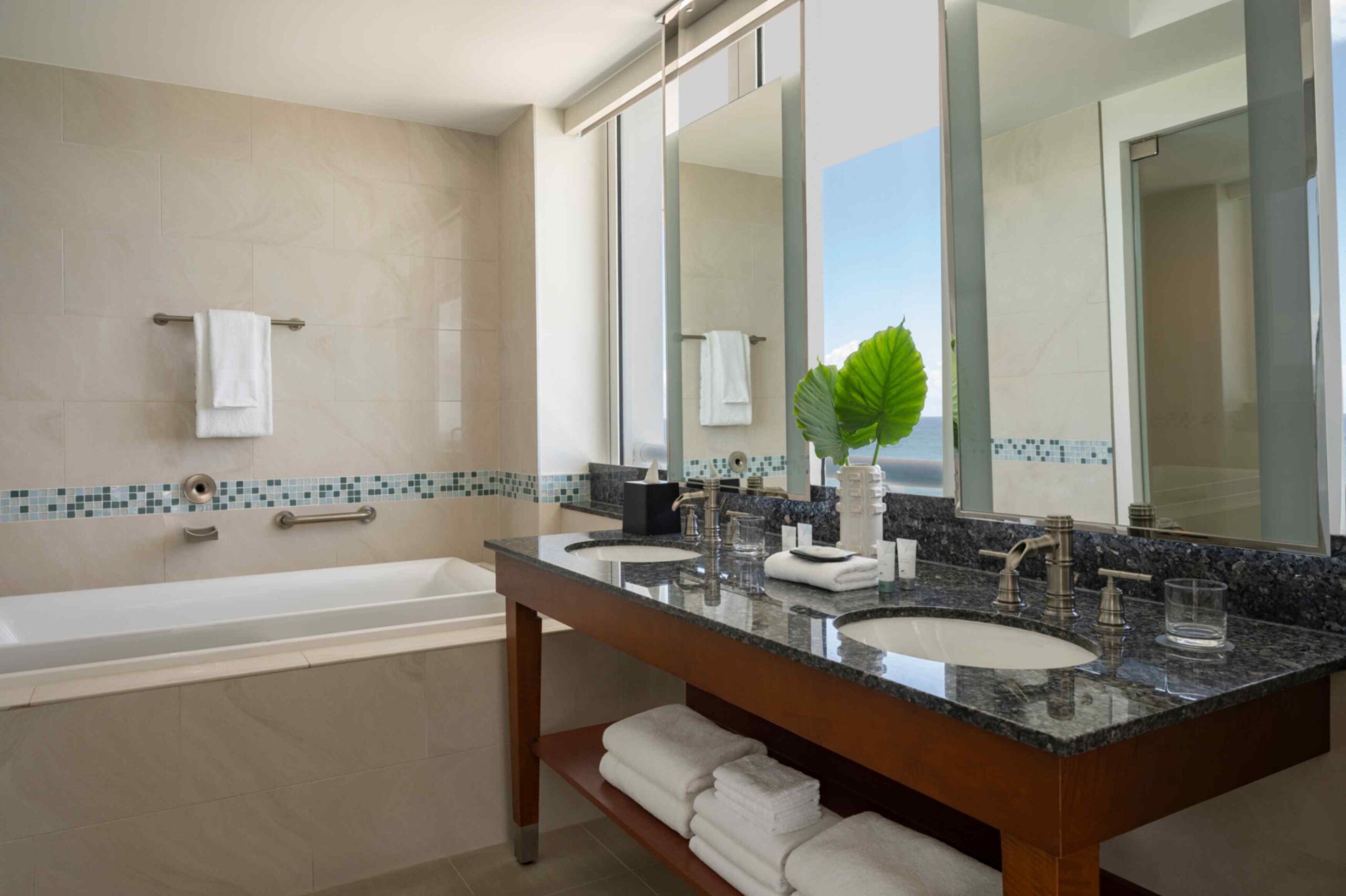A luxurious resort bathroom featuring a double vanity with dark granite countertops and two undermount sinks. Large vertical mirrors reflect light next to windows that reveal a glimpse of the ocean. On the left, a deep soaking bathtub is framed by beige wall tiles with a decorative mosaic border of blue and green tiles. White towels are neatly folded on a shelf under the vanity and hung on the wall.