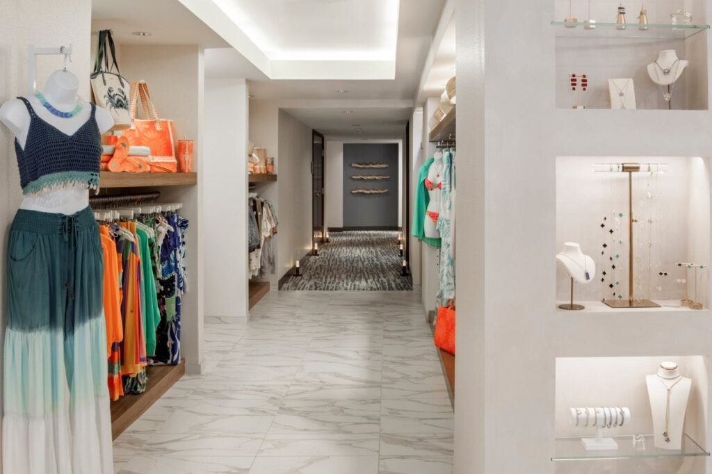 A brightly lit resort boutique corridor with white marble-patterned tile flooring. On the left, clothing racks hold vibrant orange and blue resort wear, alongside a mannequin dressed in a blue knit top and matching ombré skirt. On the right, recessed wall niches with glass shelves showcase delicate jewelry pieces, while the hallway leads down toward a grey wall with modern decor elements.