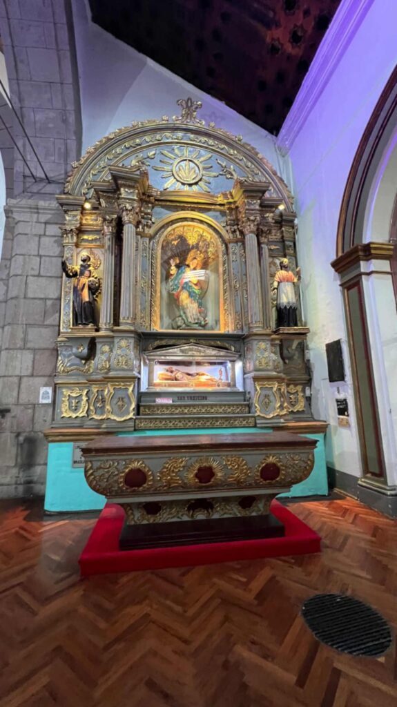 Vertical view of an ornate gold and grey baroque altar inside a Quito church featuring detailed religious statues a glass reliquary box and a matching carved wooden chest on a red platform