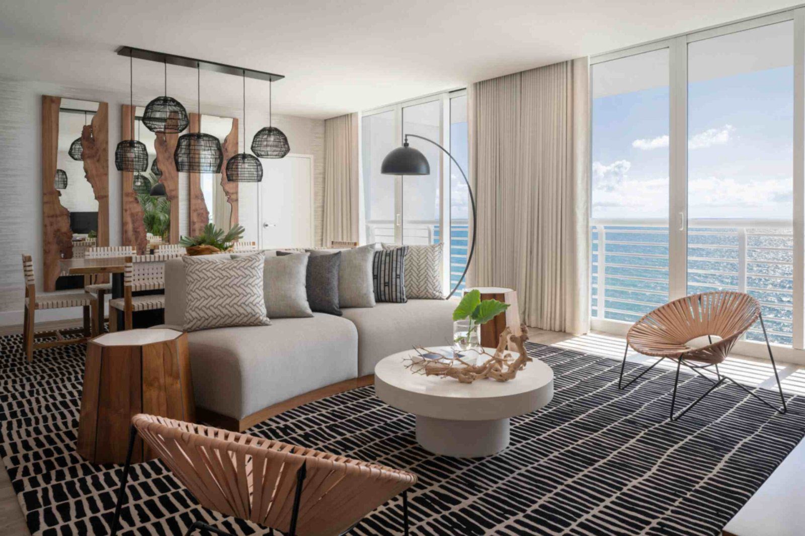 A sunlit hotel suite living room with panoramic ocean views through massive floor-to-ceiling glass doors. A large grey sectional sofa with textured throw pillows sits on a black and white patterned area rug alongside a round white coffee table and two woven accent chairs. In the background, a wooden dining table sits beneath black mesh pendant lights next to live-edge wood wall mirrors.