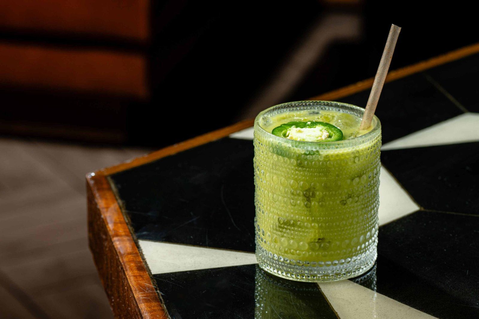 A green mezcal cocktail served in a textured, dotted glass on a black and white geometric patterned tabletop. The drink includes a paper straw and a fresh green jalapeño slice floating on top as a garnish.