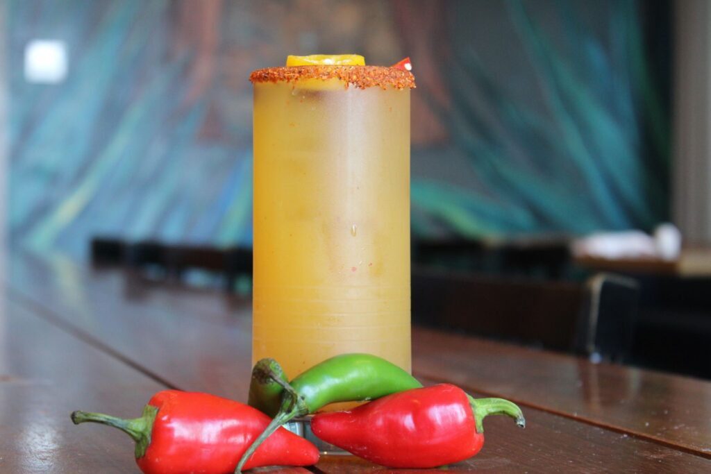 A tall iced cocktail in a frosted glass with a chili powder rim sits on a dark wood table. A yellow citrus slice floats at the top, and three whole chili peppers two red and one green rest at the base of the glass against a blurred blue background.