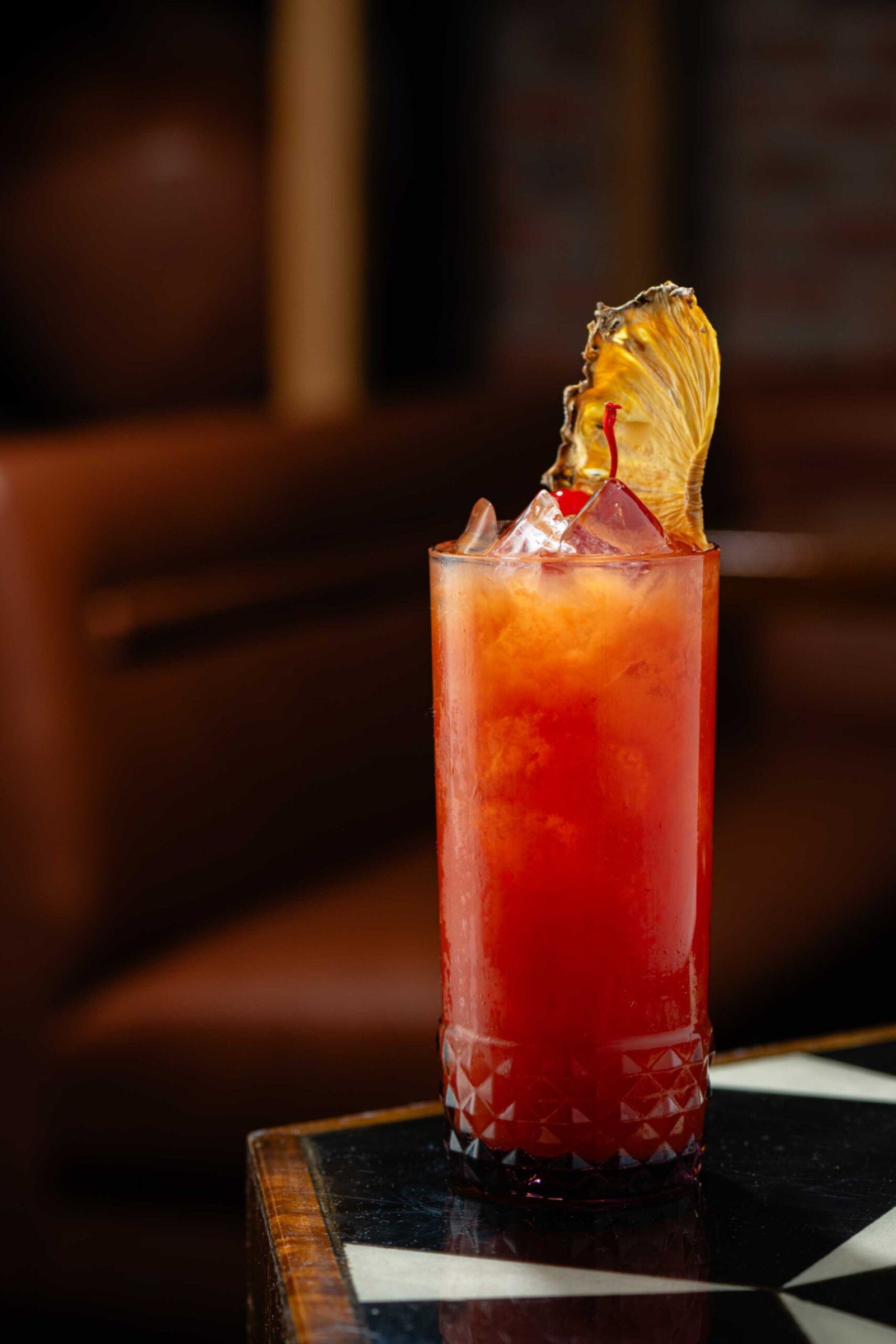 A tall vibrant red cocktail filled with crushed ice and served in a textured glass on a black and white geometric tabletop. The drink is garnished with a maraschino cherry and a delicate fan of translucent dried fruit under warm ambient lighting.