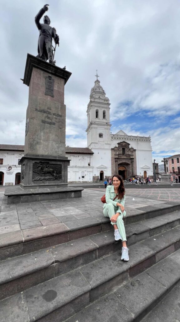 Low angle view of a woman in a light green outfit sitting on stone steps in front of the tall stone monument of Marshal Sucre and the white Baroque tower of the Santo Domingo Church in Quito Ecuador