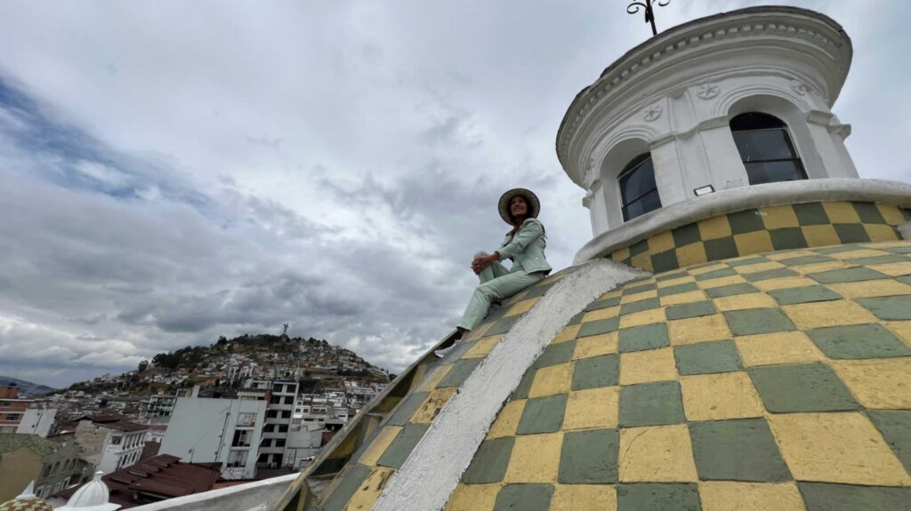 Low angle view of a woman in a light green outfit and sun hat sitting on the slope of a large yellow and green checkered dome belonging to the Quito Metropolitan Cathedral under a dramatic cloudy sky
