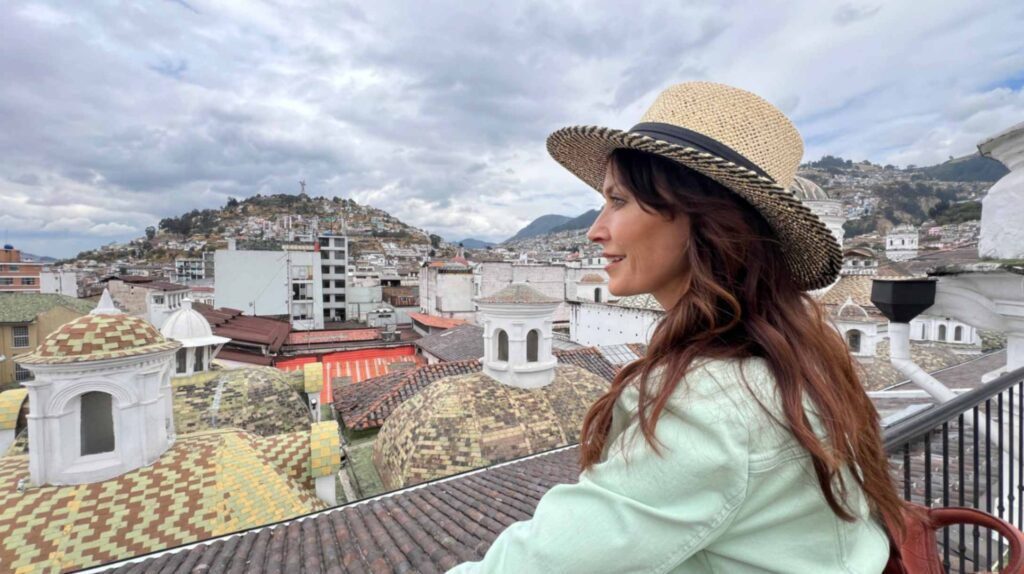 Close up side profile of a woman with long wavy brown hair wearing a woven straw hat looking out over the historic rooftops and domes of Quito Ecuador under an overcast sky