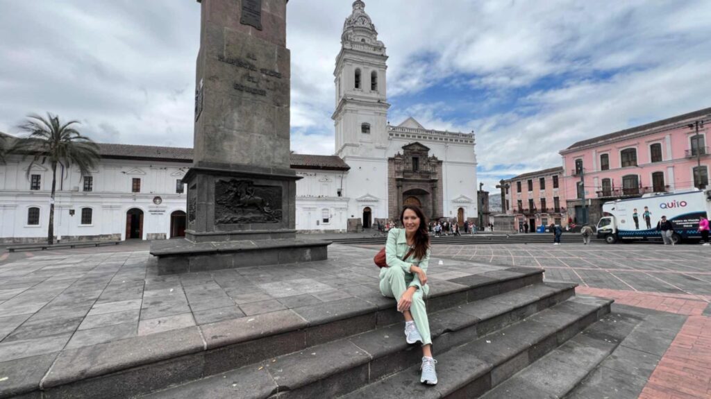 A woman in a light green outfit sitting on the stone steps of Plaza de Santo Domingo in Quito Ecuador with a massive stone monument on the left and a white colonial church tower in the background