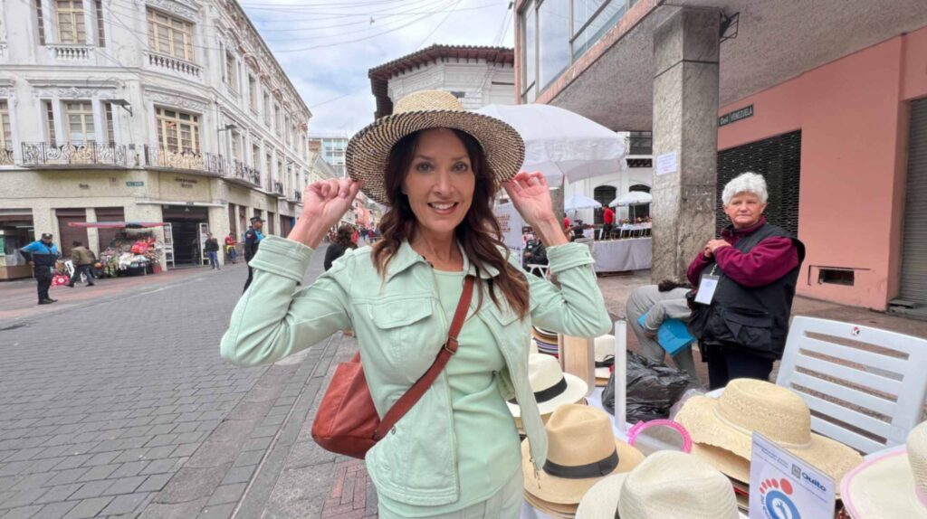 A smiling woman with long brown hair wearing a light green jacket adjusts a woven straw hat at an outdoor street vendor market booth on a cobblestone road in Quito Ecuador