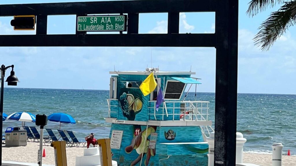 A view of Fort Lauderdale Beach through a black structural frame holding a green street sign for State Road A1A and Fort Lauderdale Beach Boulevard. On the sand sits a brightly painted blue lifeguard tower covered in sea life murals, flying yellow and purple warning flags. Blue beach umbrellas and lounge chairs line the shore in the background beneath a clear blue sky.